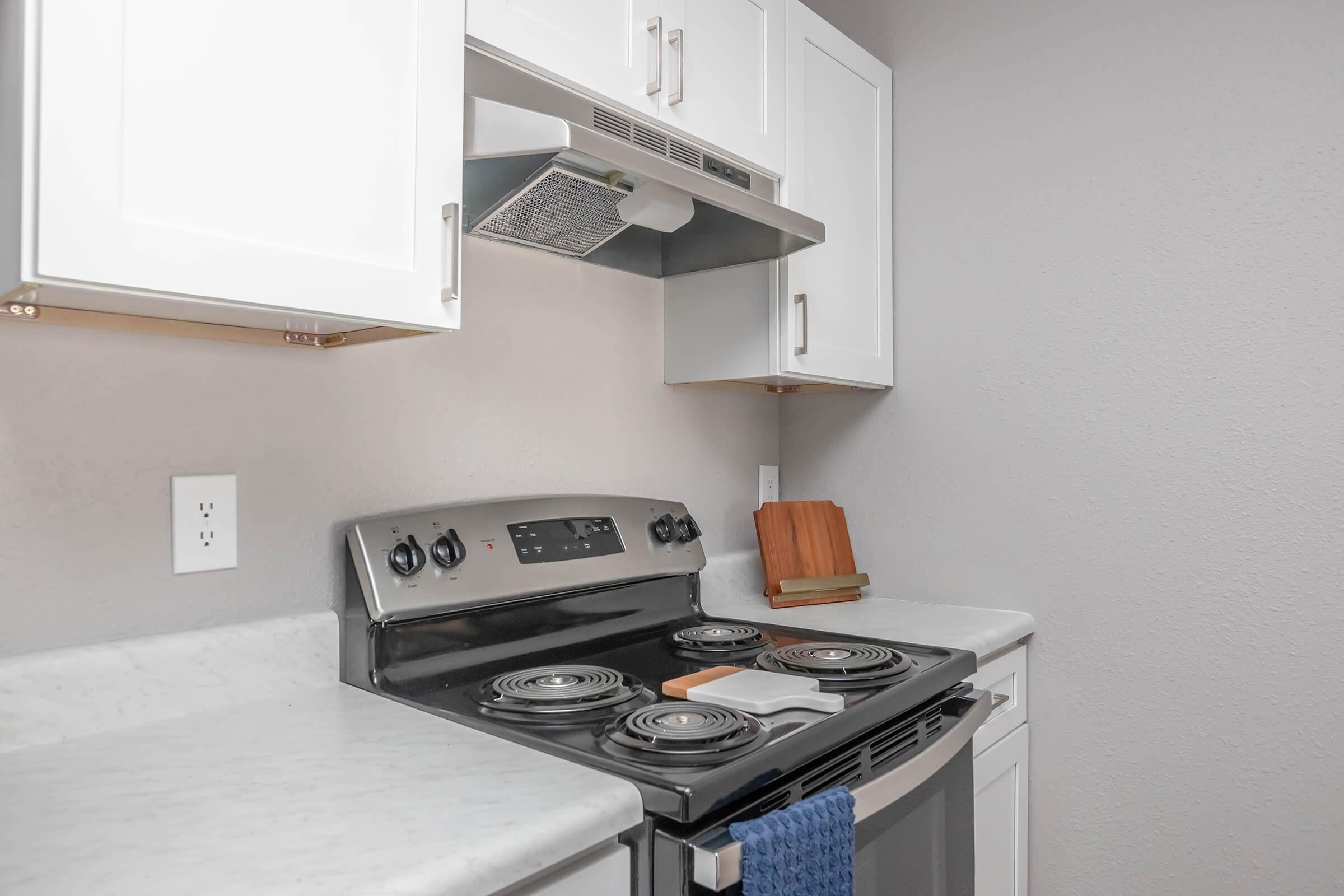 A modern kitchen with white cabinets, featuring a stainless steel electric stove with four burners, a silver range hood, and a marble countertop. A wooden cutting board is placed on the counter next to the stove, which is equipped with a blue dish towel hanging from the front.