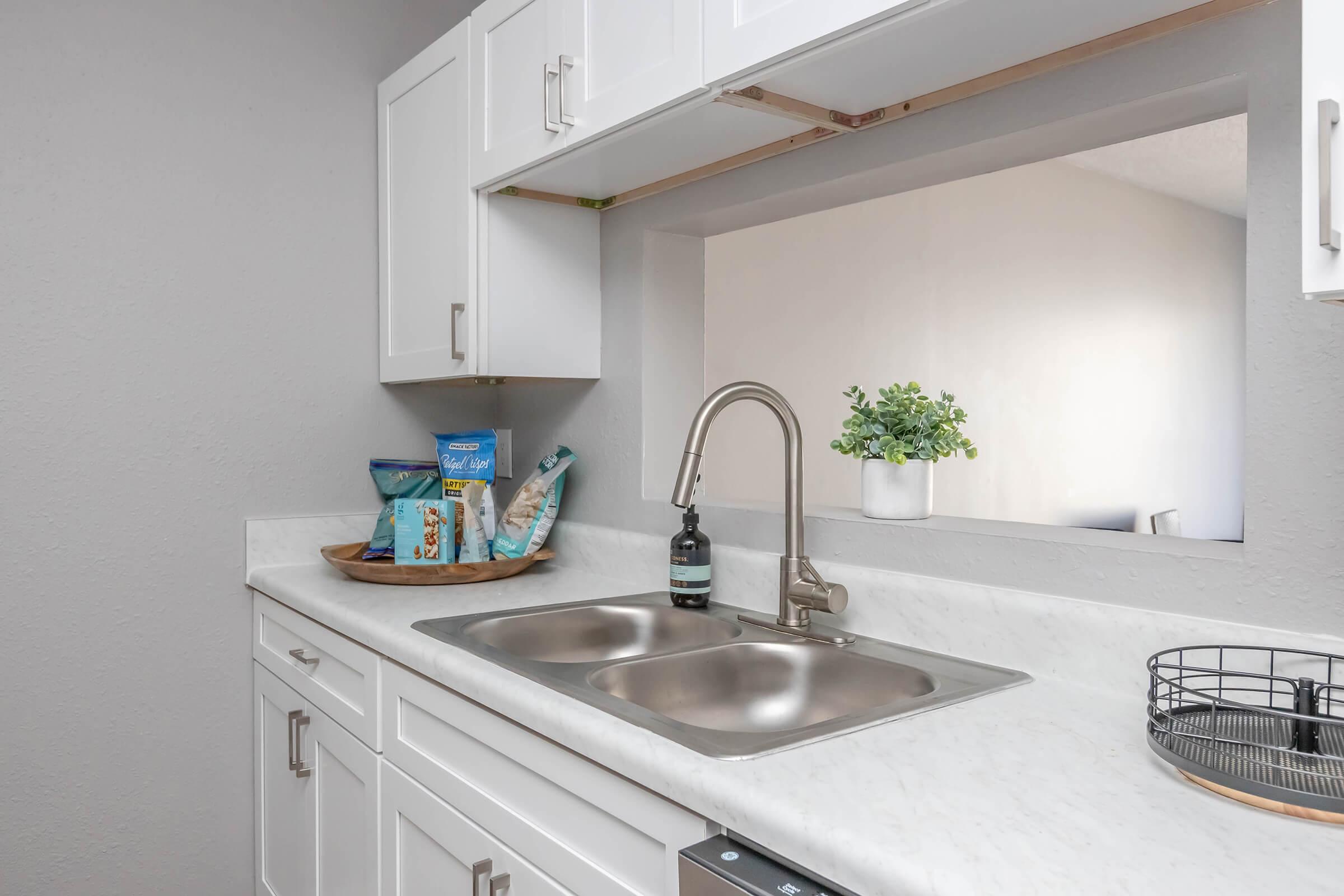 A modern kitchen with white cabinetry and a stainless steel sink. There are bags of snacks on a wooden tray, a potted plant, and a bottle of dish soap near the sink. A small wire basket sits on the counter, and a mirror reflects light from the adjacent room. The walls are painted a light gray.