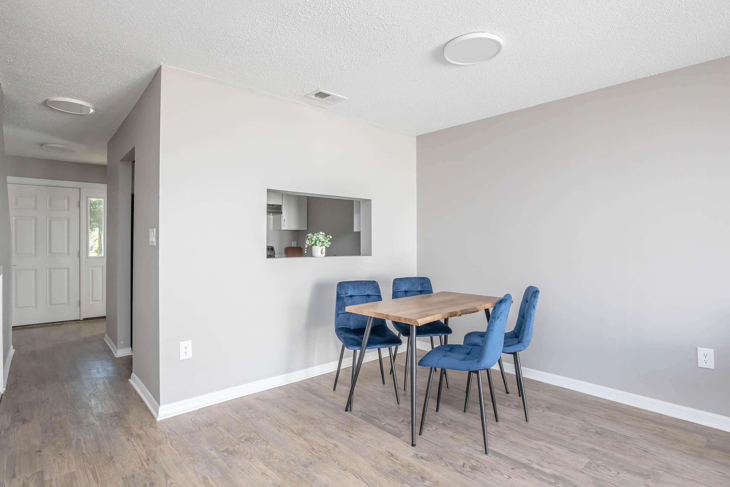 A bright, modern dining area featuring a simple wooden table with four blue upholstered chairs. The walls are light gray, and there is an opening leading to a kitchen area. A small indoor plant decorates the table, and there's a white door visible in the hallway. The floor is light wood laminate.