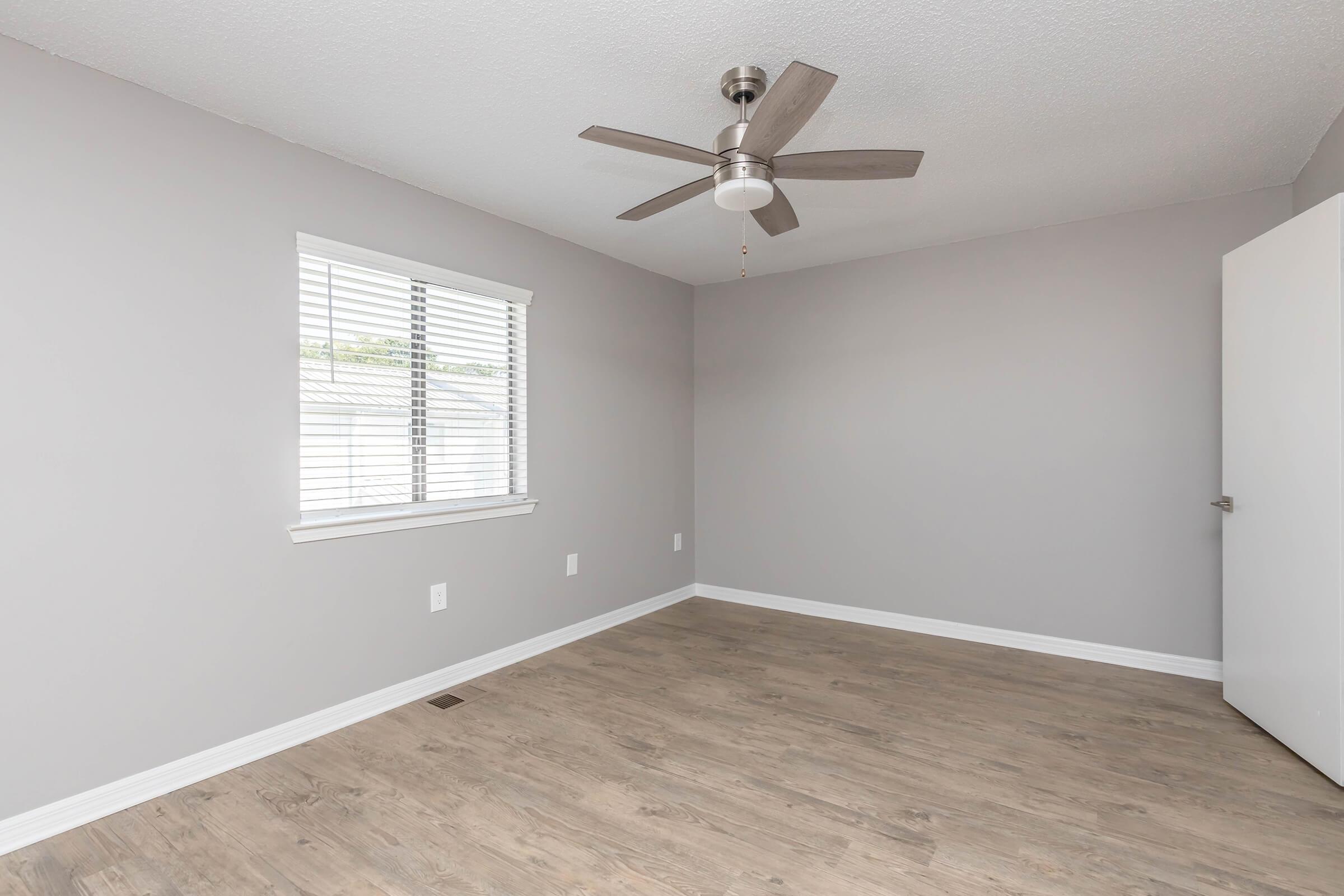 A well-lit, empty room with light gray walls and a ceiling fan. There is a window with blinds on the left side and a white door partially visible on the right. The floor is made of light-colored wood, creating a clean and minimalist aesthetic.