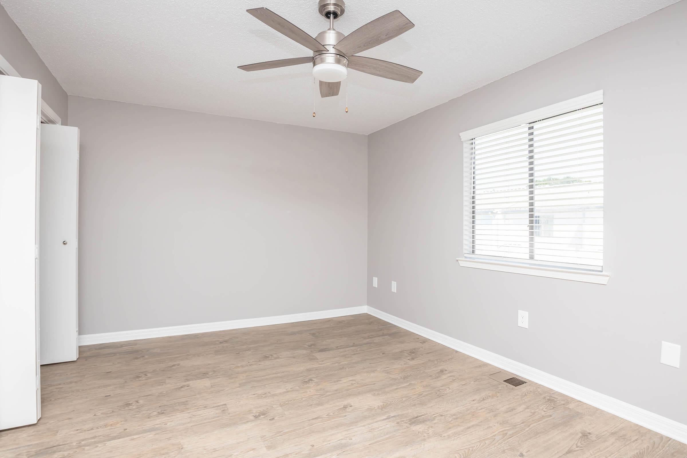 A minimalistic empty room featuring light gray walls, a ceiling fan with a light fixture, and a window adorned with blinds. The floor is made of light wood, and there is a white closet on one wall. The room has a clean and modern aesthetic, suitable for various uses.