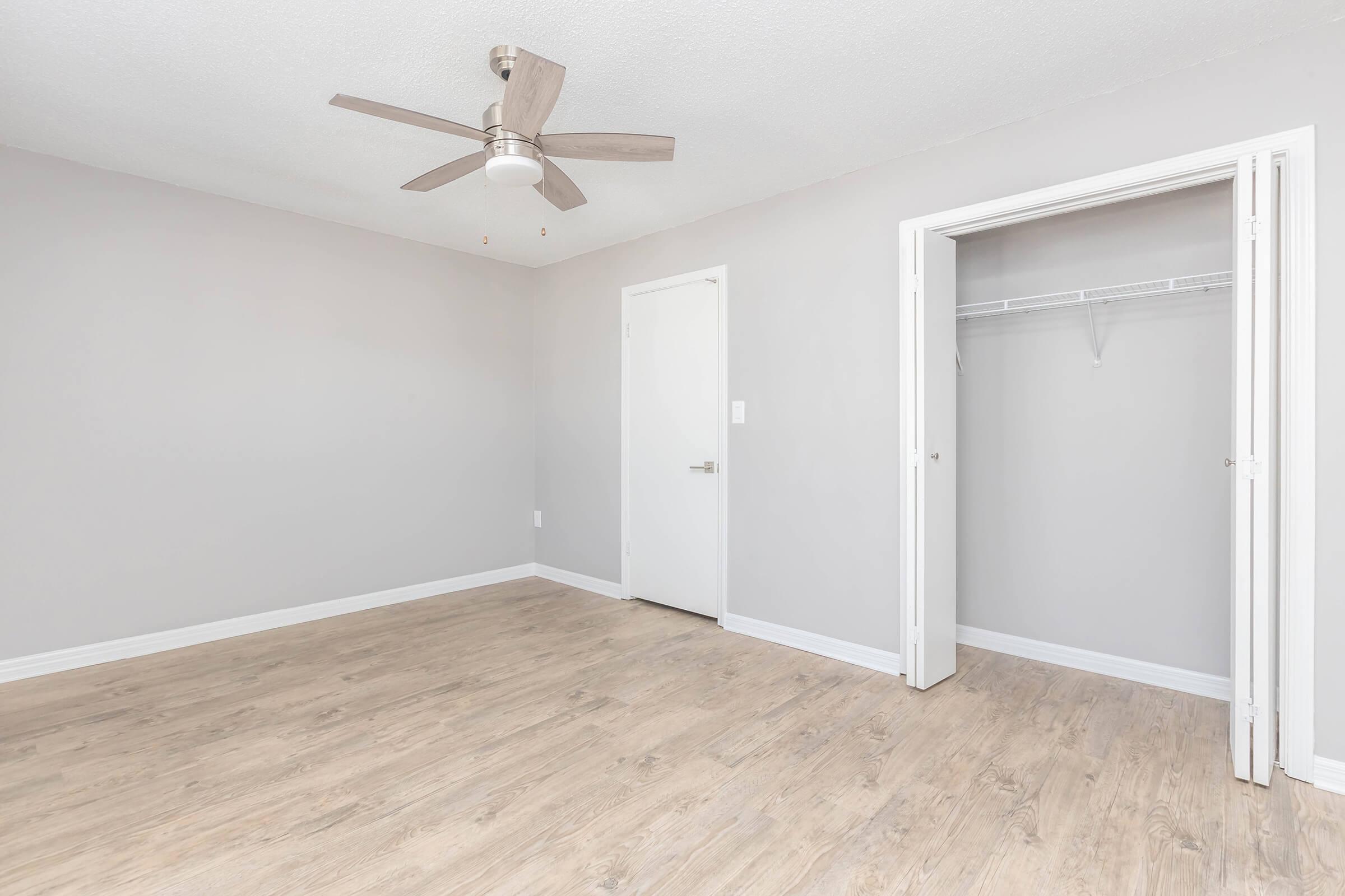 Empty room with light gray walls and wooden flooring. A ceiling fan is mounted in the center, and there is a white door leading to a closet. The space appears bright and minimalistic, suitable for various uses or decoration.