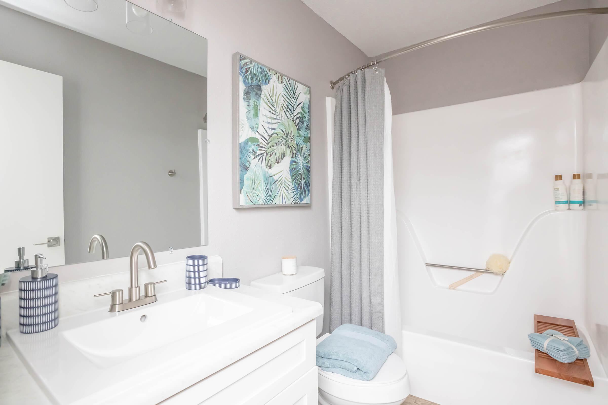 A modern bathroom featuring a white sink with a silver faucet, a large mirror, light gray walls, and a bathtub with a shower curtain. There is a decorative wall art piece with a tropical theme, a small wooden shelf with toiletries, and blue towels displayed neatly.