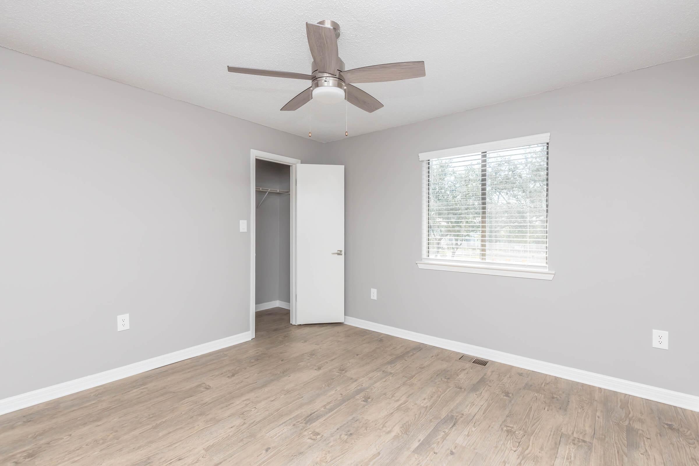 Empty room with light gray walls and a ceiling fan. A window with horizontal blinds allows natural light in, and a closet door is slightly ajar, revealing a minimal closet space. The floor is wooden laminate, and the overall atmosphere is bright and spacious.