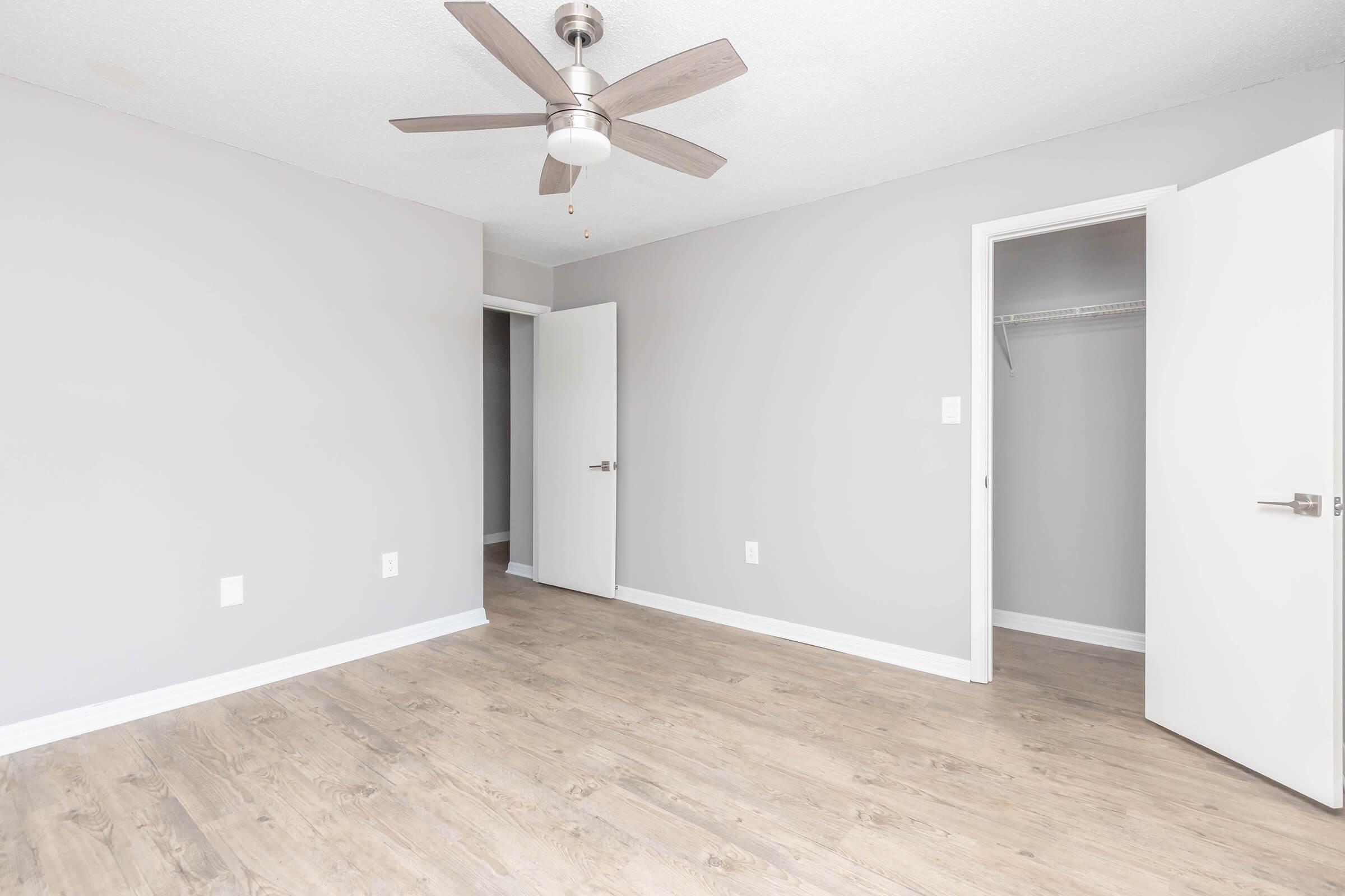 A minimalistic room with gray walls and light wood flooring. A ceiling fan is mounted in the center. There are two white doors: one open revealing a closet space, and the other closed. The room is well-lit and spacious, providing a clean and modern aesthetic.