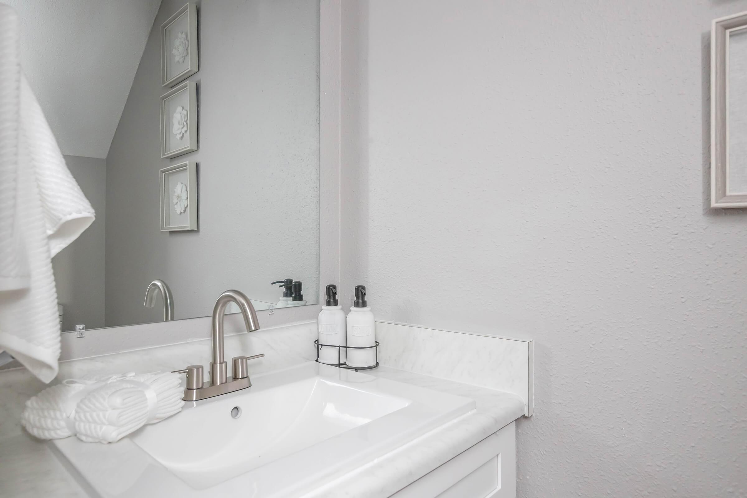 A clean bathroom sink area featuring a modern faucet, white soap dispenser, and a decorative towel hanging nearby. A mirror reflects the space, and framed floral artwork is visible on the wall, adding a touch of elegance to the neutral-colored decor.