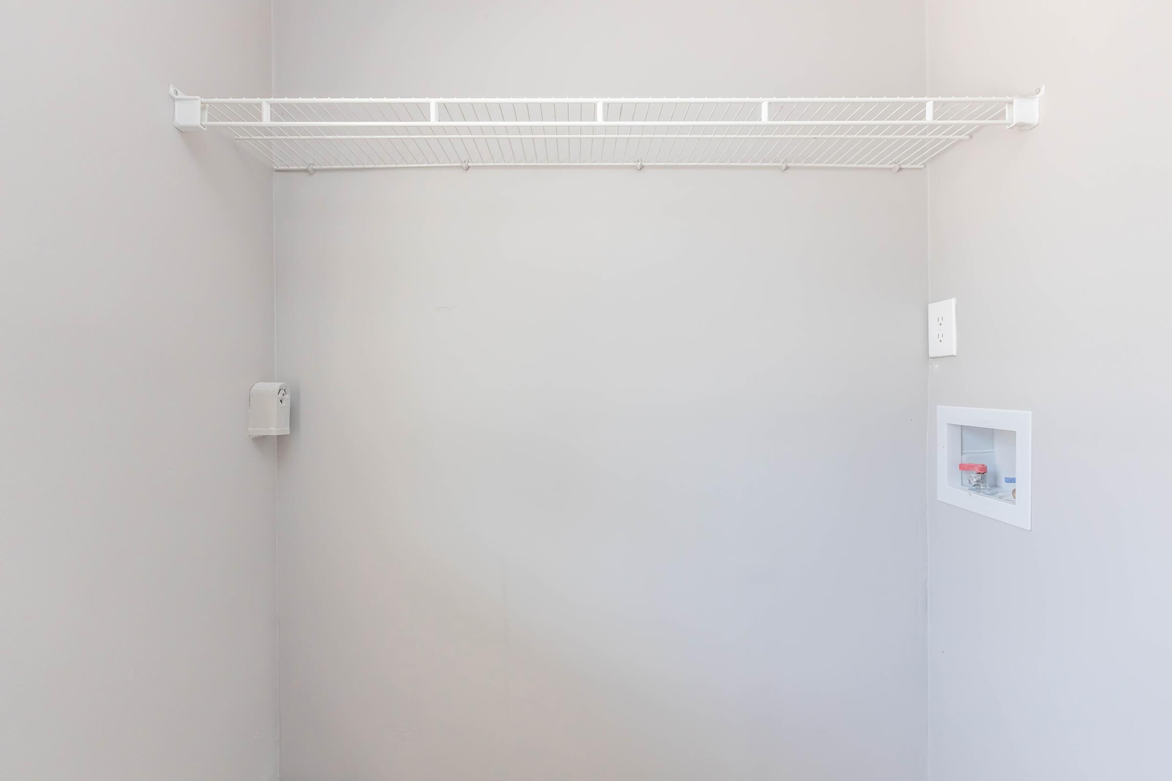 A minimalistic, empty room with light gray walls. There is a white wire shelf mounted on the wall, and a small storage nook featuring a white frame with a red item inside. The overall space appears clean and uncluttered, suggesting it may be a utility or storage area.
