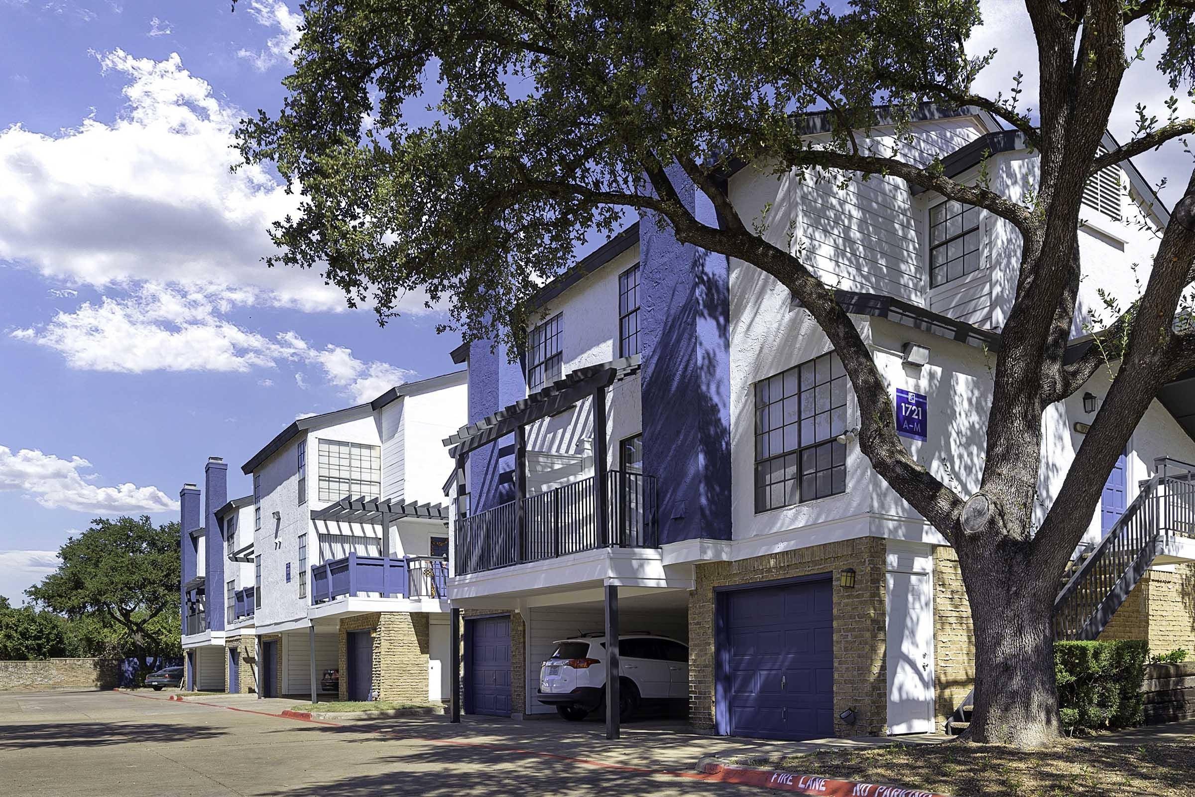 A residential building with modern architecture featuring two adjacent units, each with a garage. The façade includes blue accents and large windows, complemented by a shaded outdoor area. A well-maintained tree stands in the foreground, and the sky is bright with scattered clouds.
