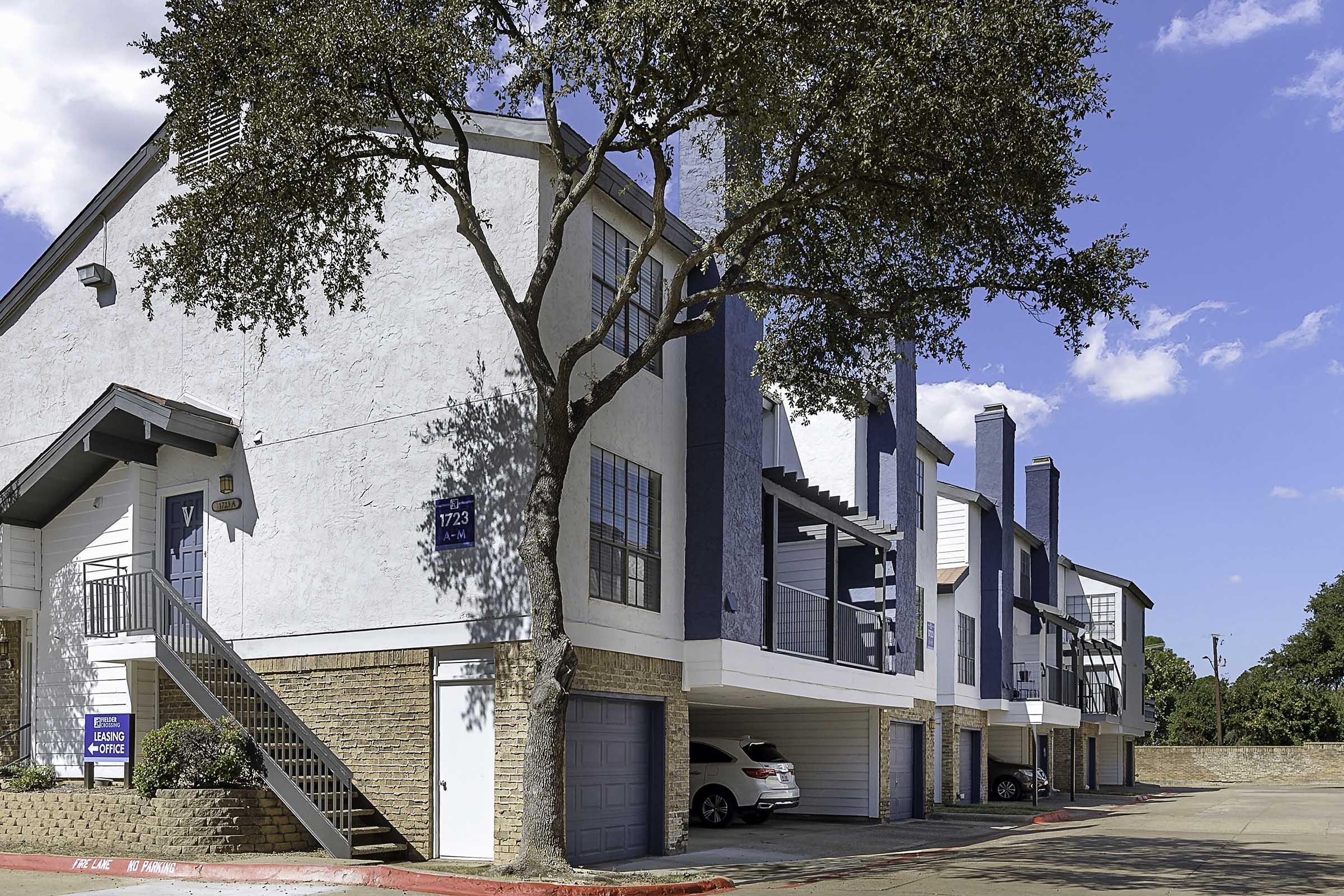 A view of a multi-story apartment building with a combination of white and blue exteriors. The building features multiple garages and a staircase leading to the upper units. There are parked cars in the garage area and a clear blue sky in the background. Trees provide some greenery around the property.