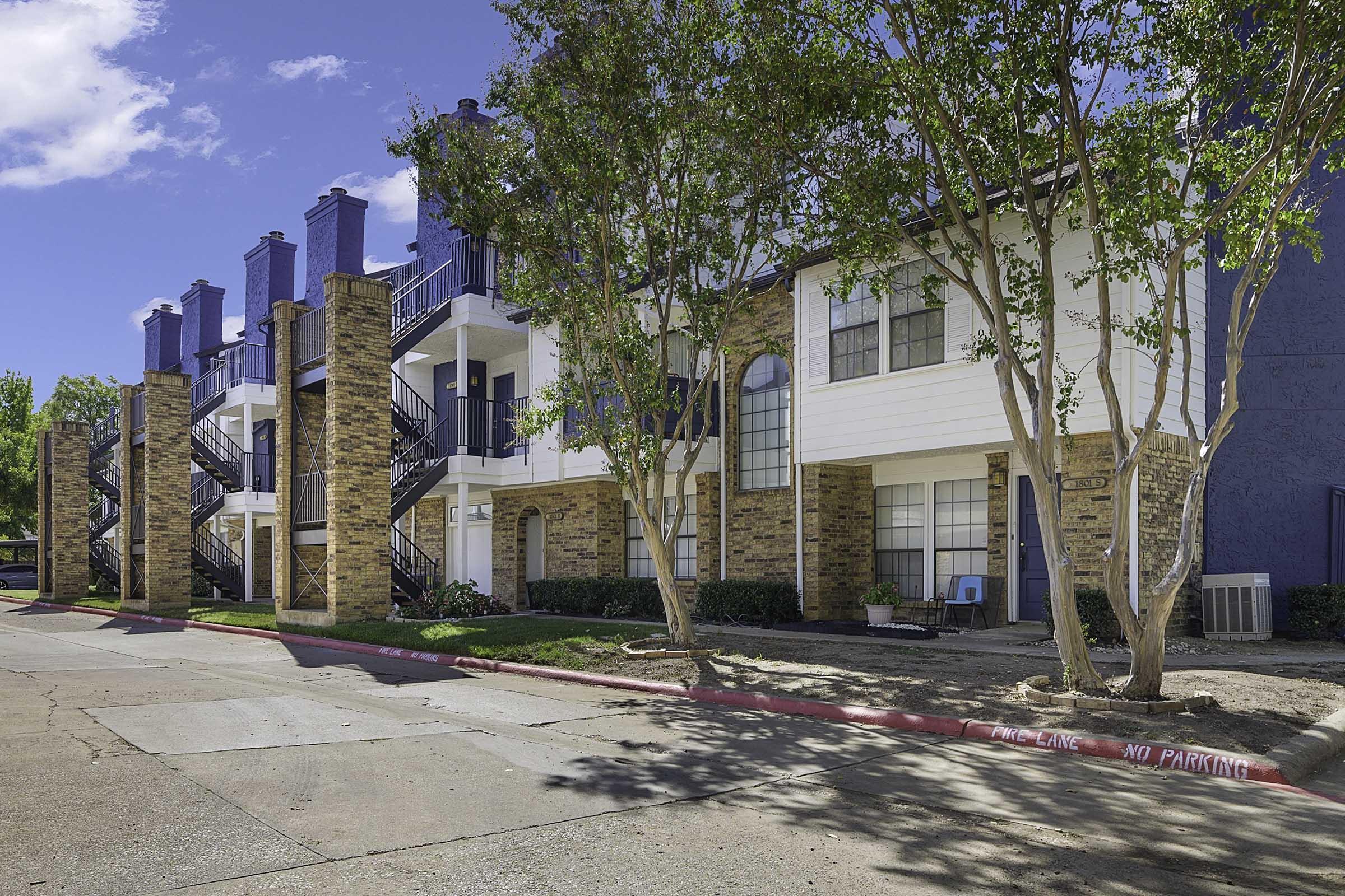 A modern apartment complex featuring multiple two-story buildings. The exterior showcases a combination of brick and wood siding, with decorative balconies and staircases. Lush trees provide greenery in the foreground, and a clear blue sky is visible above. A no parking zone is marked on the street.