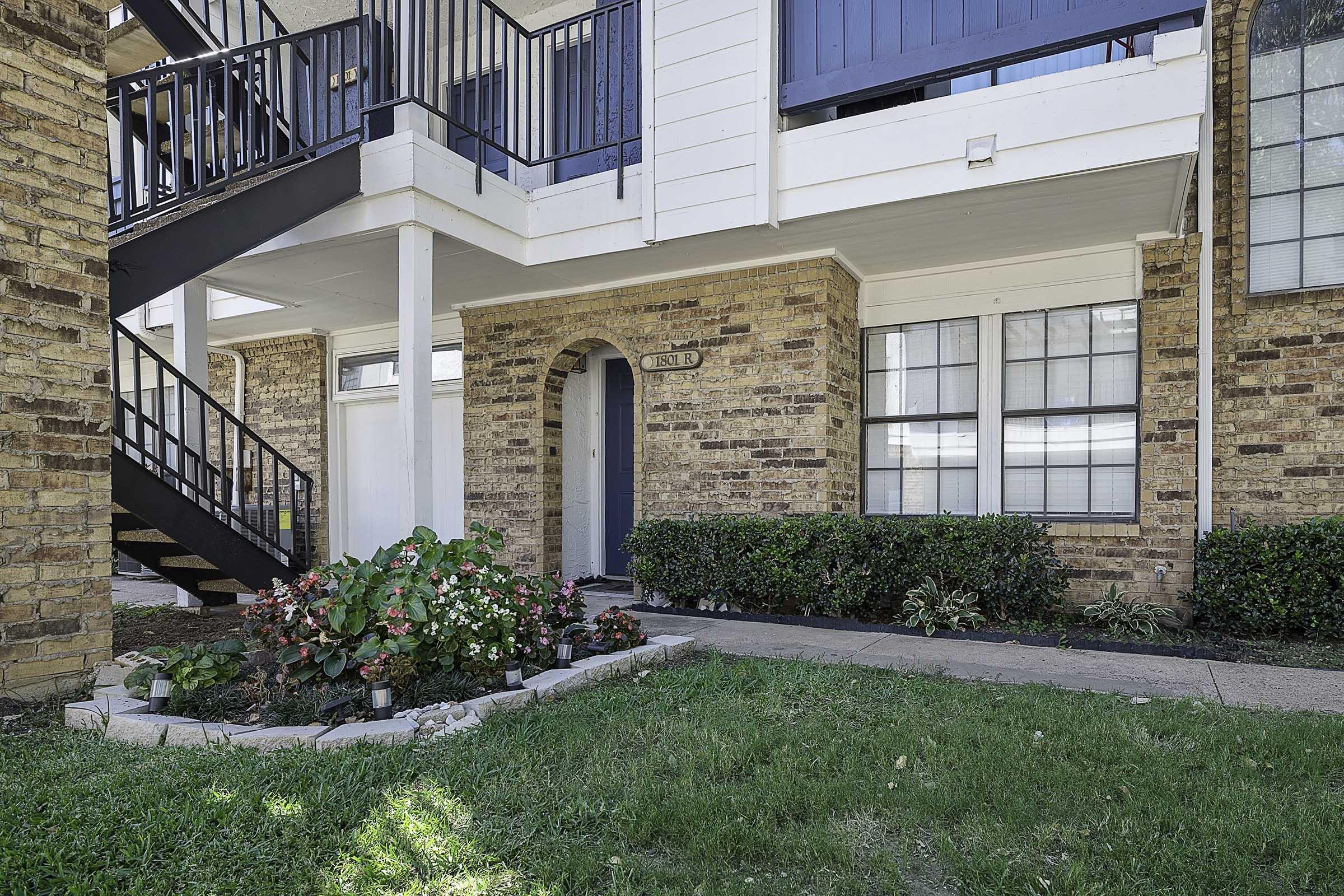 Exterior view of an apartment building featuring a brick facade, a set of black metal stairs leading to the upper floor, and a small garden with colorful flowers in the foreground. The entrance door is painted blue, and several windows can be seen on the ground level. Green lawn surrounds the pathway.