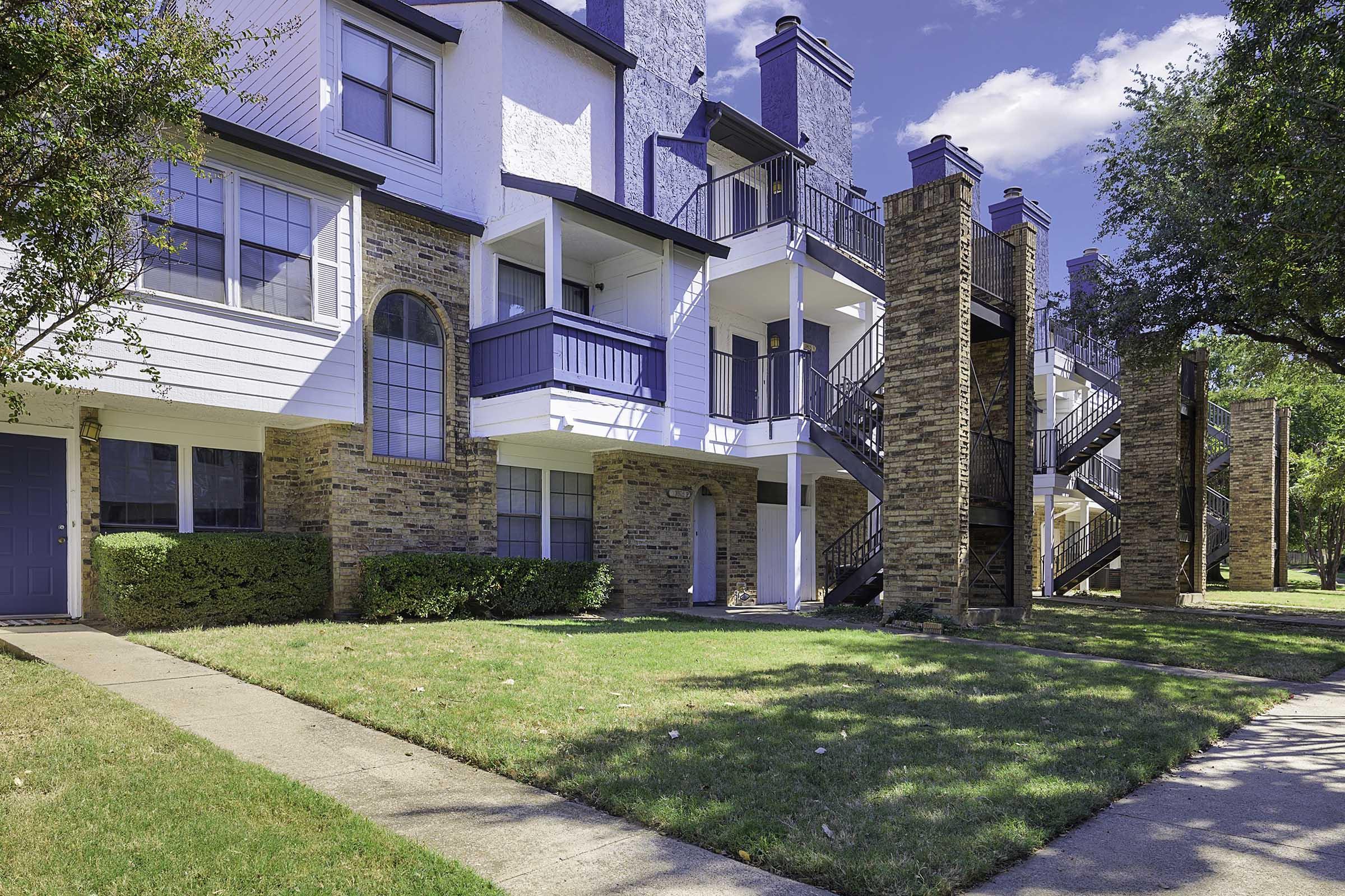 Three-story apartment building with a mix of brick and white siding. Balconies are present on each floor, with black railings. Lush green grass is maintained in front of the building, and there are trees nearby. The sky is clear with a few clouds, creating a bright atmosphere.