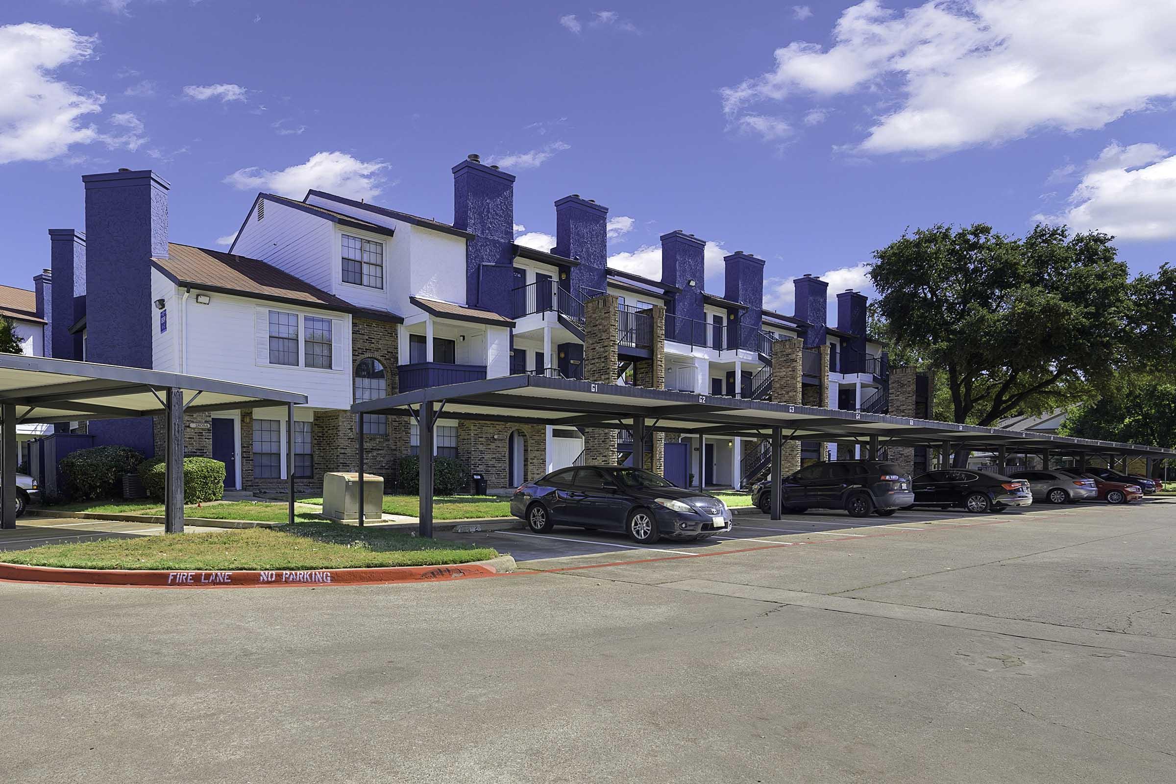 A multi-unit apartment building featuring a combination of blue and white exteriors, with stone accents. The building has several covered parking spaces in front, and lush green landscaping surrounds the property. The sky is clear with a few clouds.