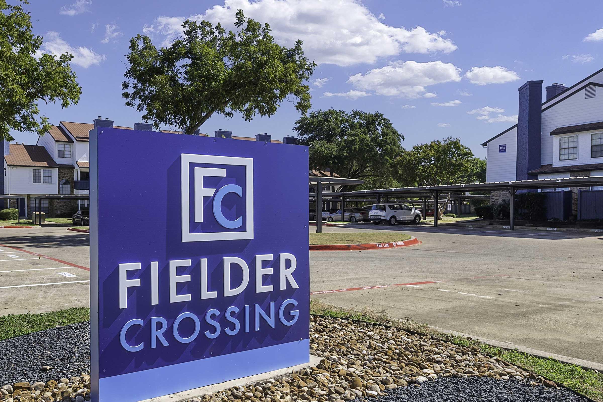 A blue sign featuring the text "Fielder Crossing" and "FC" prominently displayed, set in a landscaped area with gravel and trees. In the background, there are residential buildings and a parking area under a clear blue sky with scattered clouds.