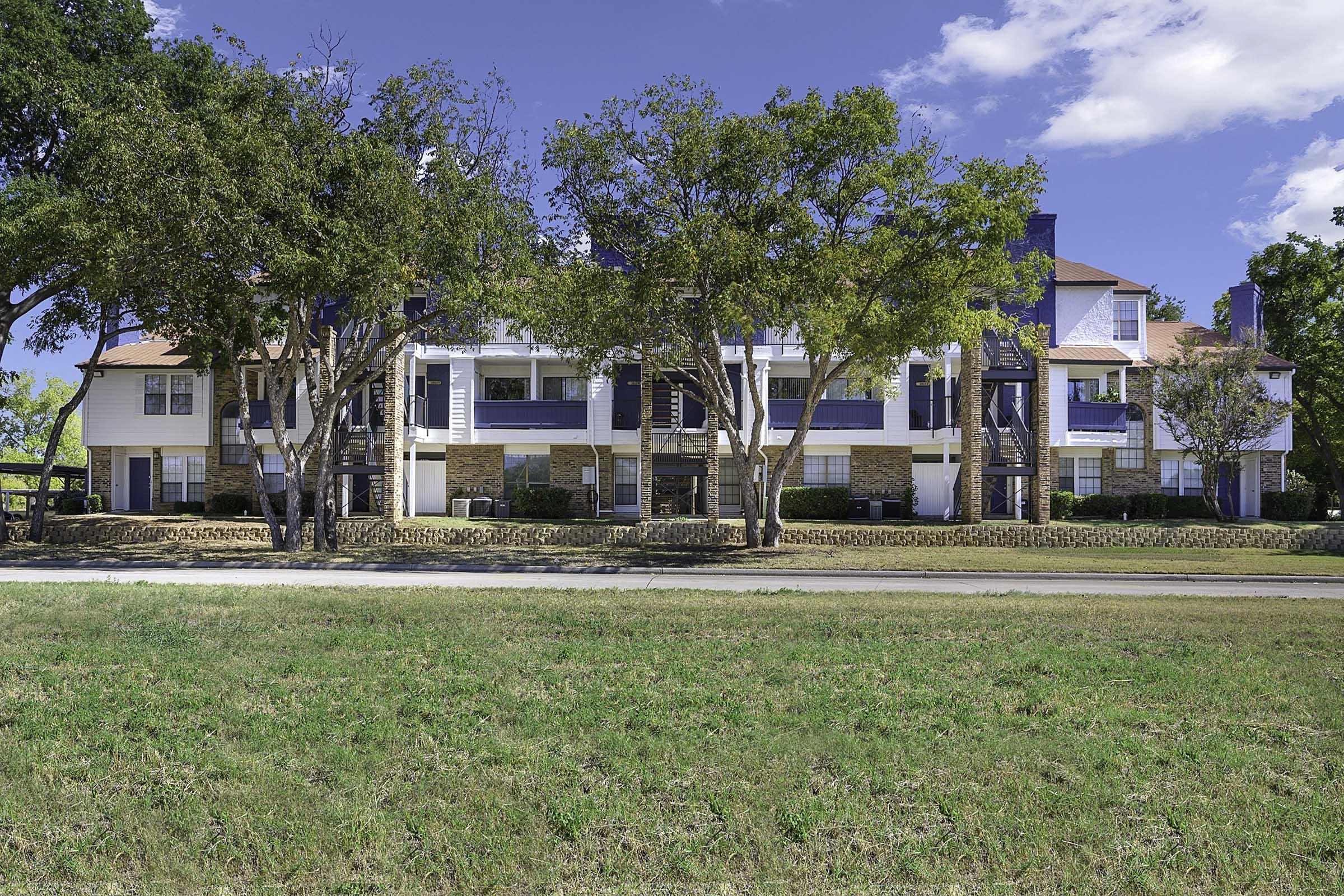 A two-story residential building with blue and white facade, featuring several balconies and large windows. Surrounded by lush green grass and a few trees, the scene is set against a clear blue sky with a few clouds.