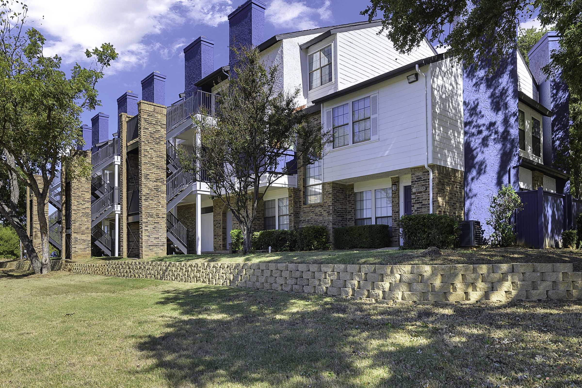 A multi-story apartment building featuring a mix of brick and white siding. It has three levels with balconies, surrounded by green lawns and trees. A stone retaining wall runs along the front of the property, with blue sky and clouds visible in the background.