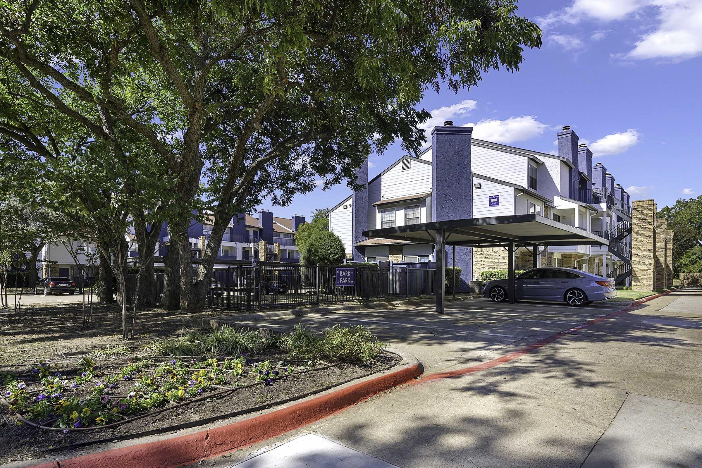 A view of an apartment complex featuring two-story buildings with blue roofs. The parking area includes a gray car and landscaped flower beds with colorful blooms. There are several trees providing shade, and the sky is clear with a few clouds. A sign for "Tamarack Square" is visible.