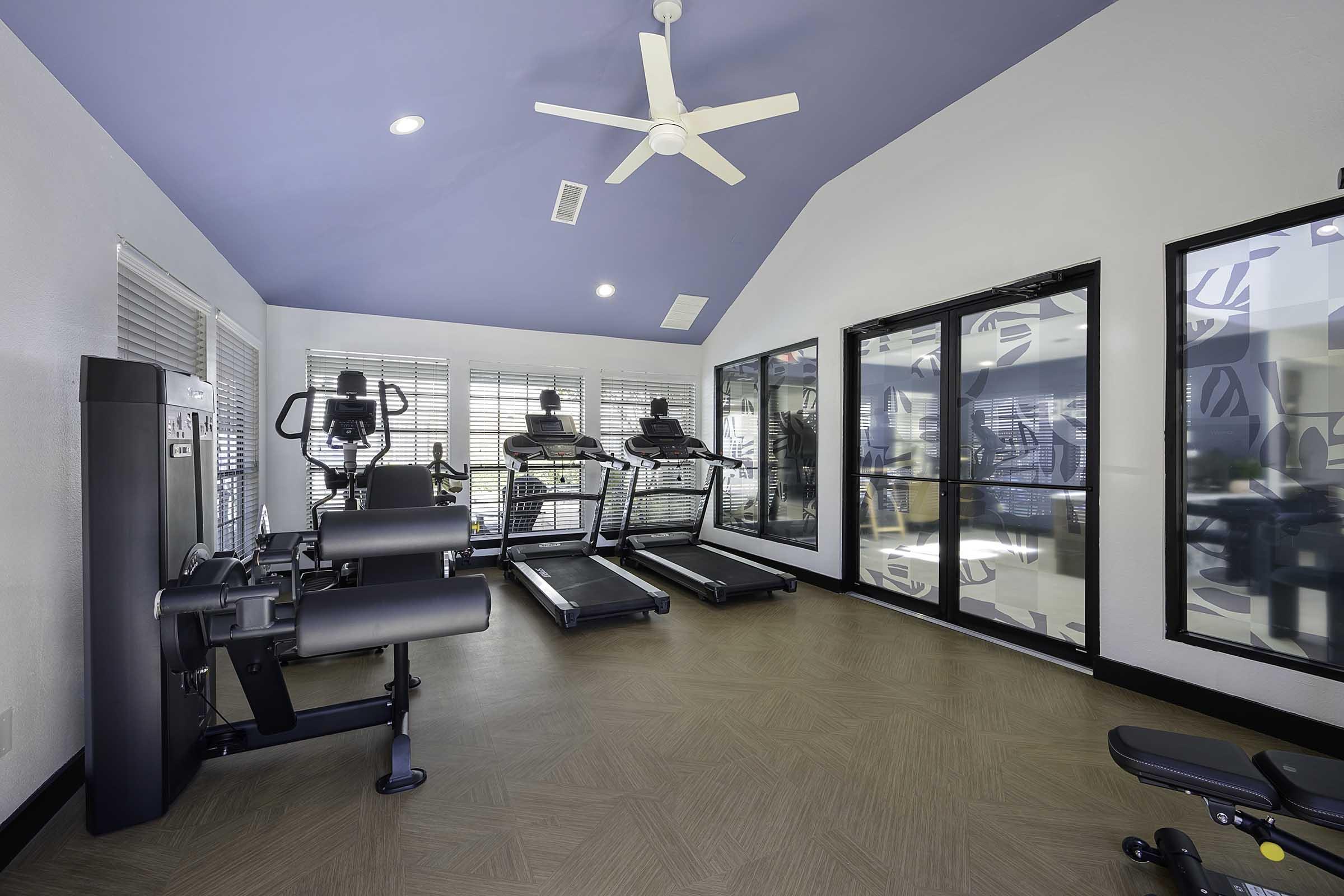 A modern gym interior featuring several exercise machines, including treadmills and stationary bikes. The room has large windows with blinds, a ceiling fan, and light blue walls, creating a bright and airy atmosphere. Fitness equipment is neatly arranged, emphasizing a clean and inviting workout space.