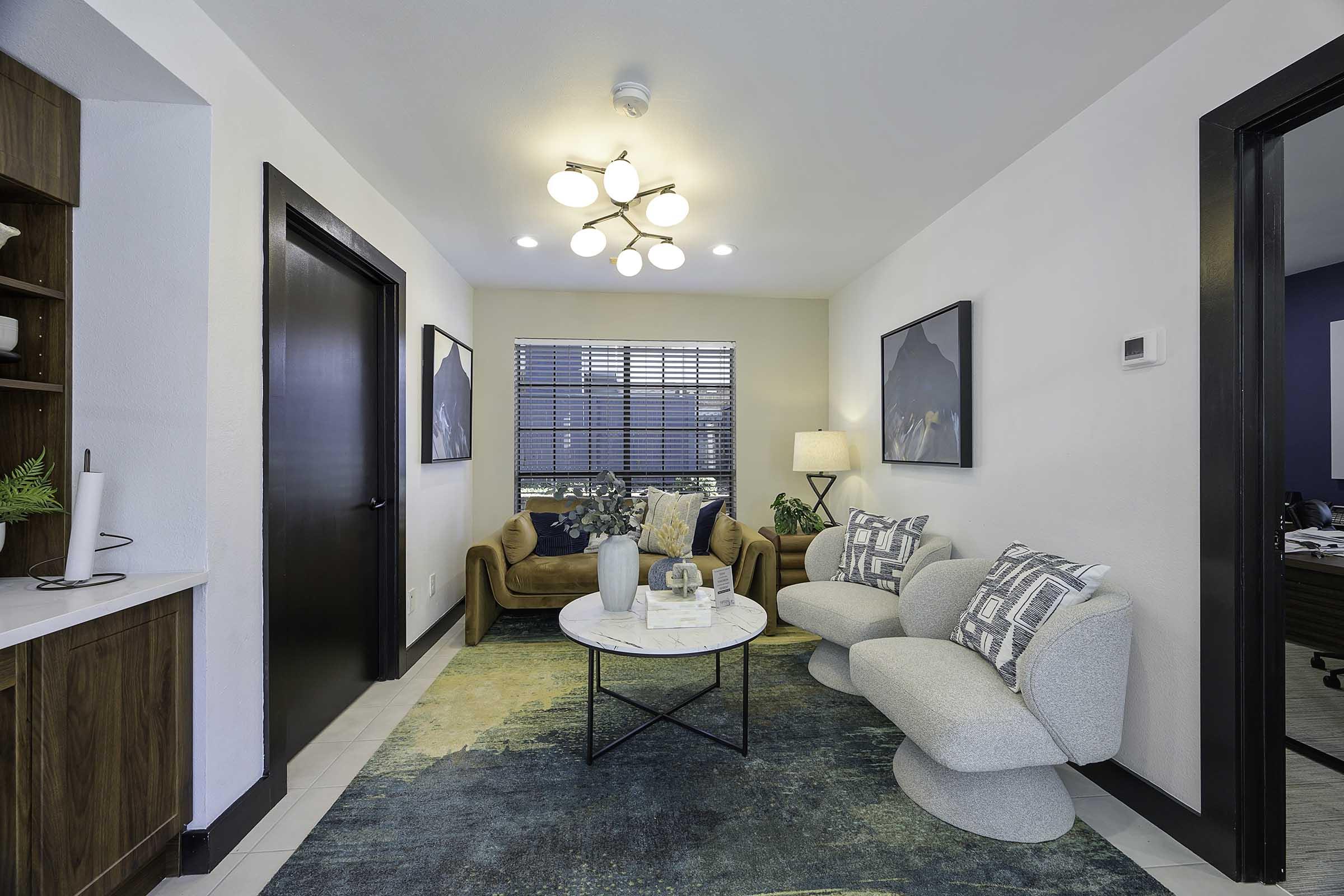 A modern living room featuring a light-colored couch, two plush chairs, a circular coffee table, and a decorative rug. There are two framed artworks on the walls, a window with blind treatment, and a small bookshelf with greenery. The room is bright and inviting, with warm lighting.