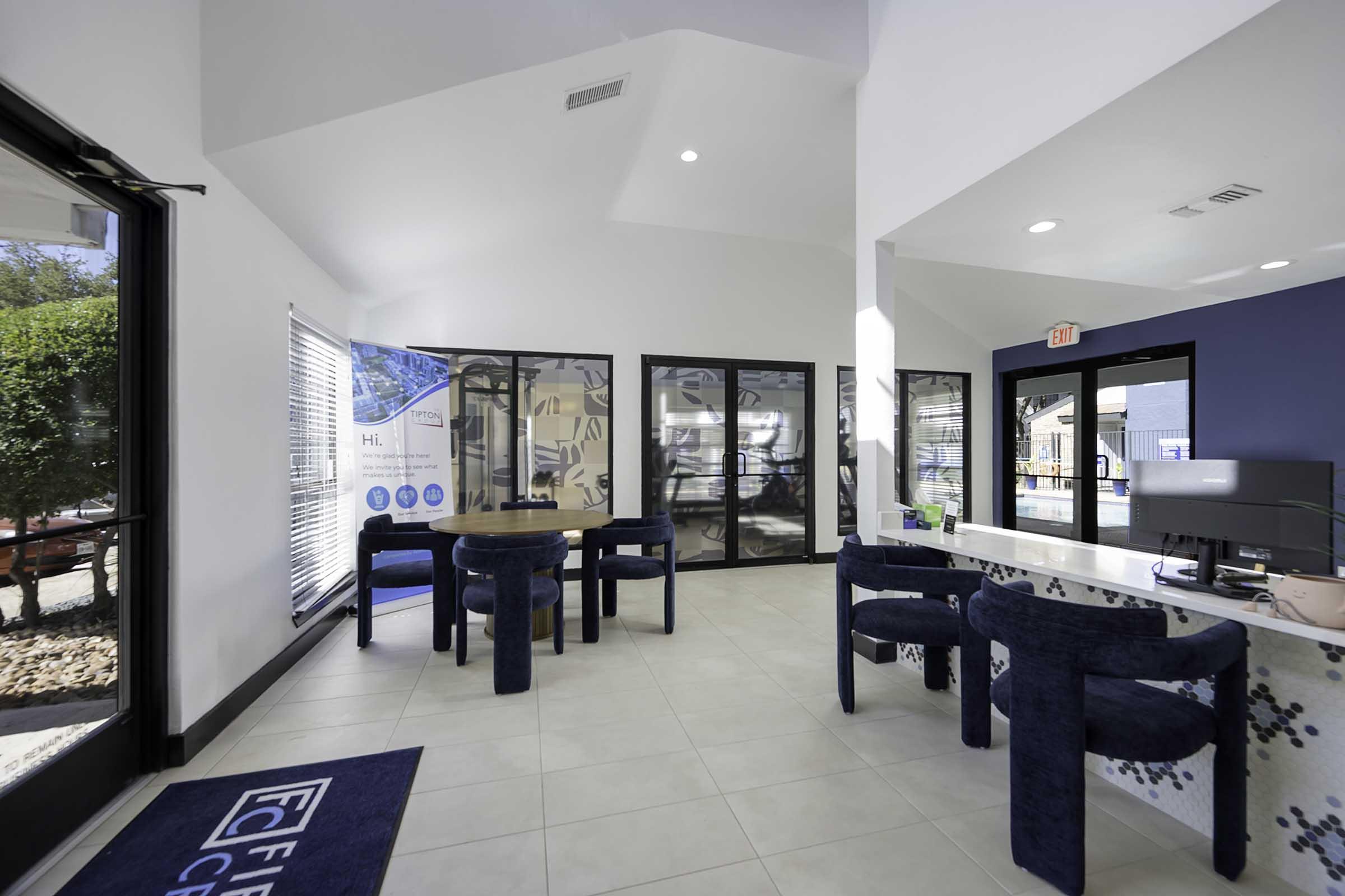 Interior of a modern office space featuring a round wooden table surrounded by blue upholstered chairs. A reception area with a desk and a computer is visible, alongside large glass windows allowing natural light. Walls are painted in light colors with decorative elements.