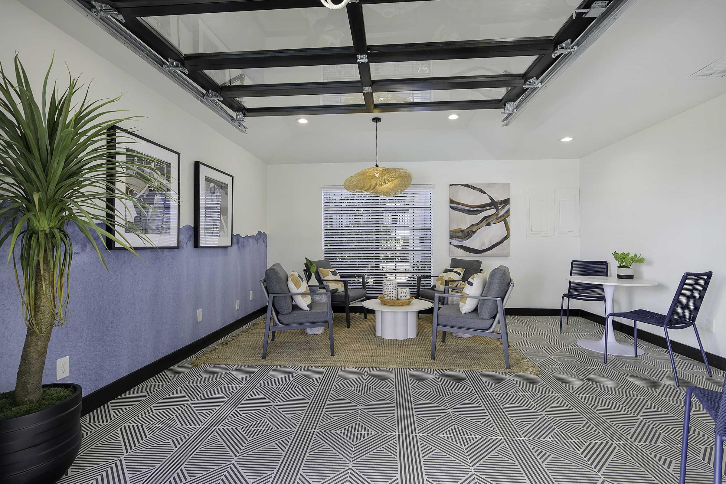 A modern lounge area featuring two gray armchairs and a round white table, surrounded by patterned tiles. A decorative plant is in one corner, while the walls showcase framed black-and-white art. Natural light enters through large windows, complemented by a stylish pendant light overhead.