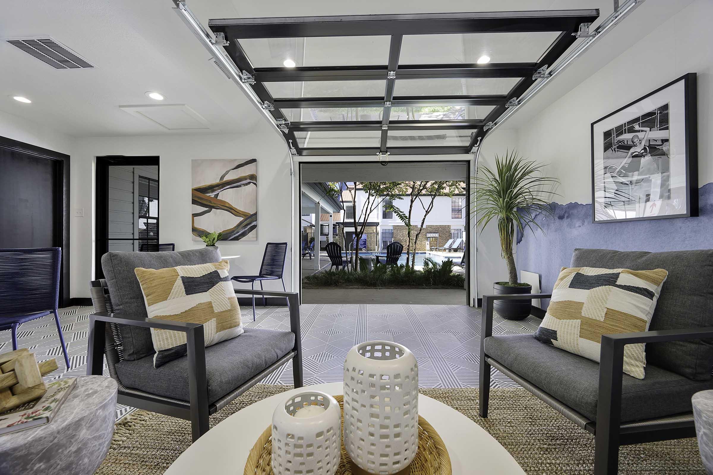 Modern interior living space featuring two comfortable gray armchairs with decorative pillows, a round coffee table with white candle holders, and large glass doors that open to an outdoor area with a view of trees and a pool. Natural light illuminates the room, which has a stylish and contemporary design.
