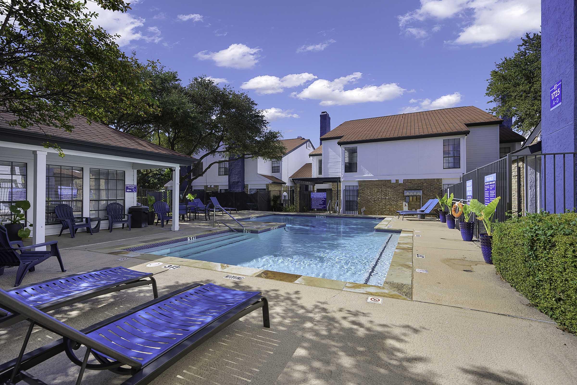 A clear blue swimming pool surrounded by lounge chairs, potted plants, and trees. In the background, there are two residential buildings with white and light-colored exteriors. The sky is bright with a few clouds, creating a cheerful and inviting atmosphere.