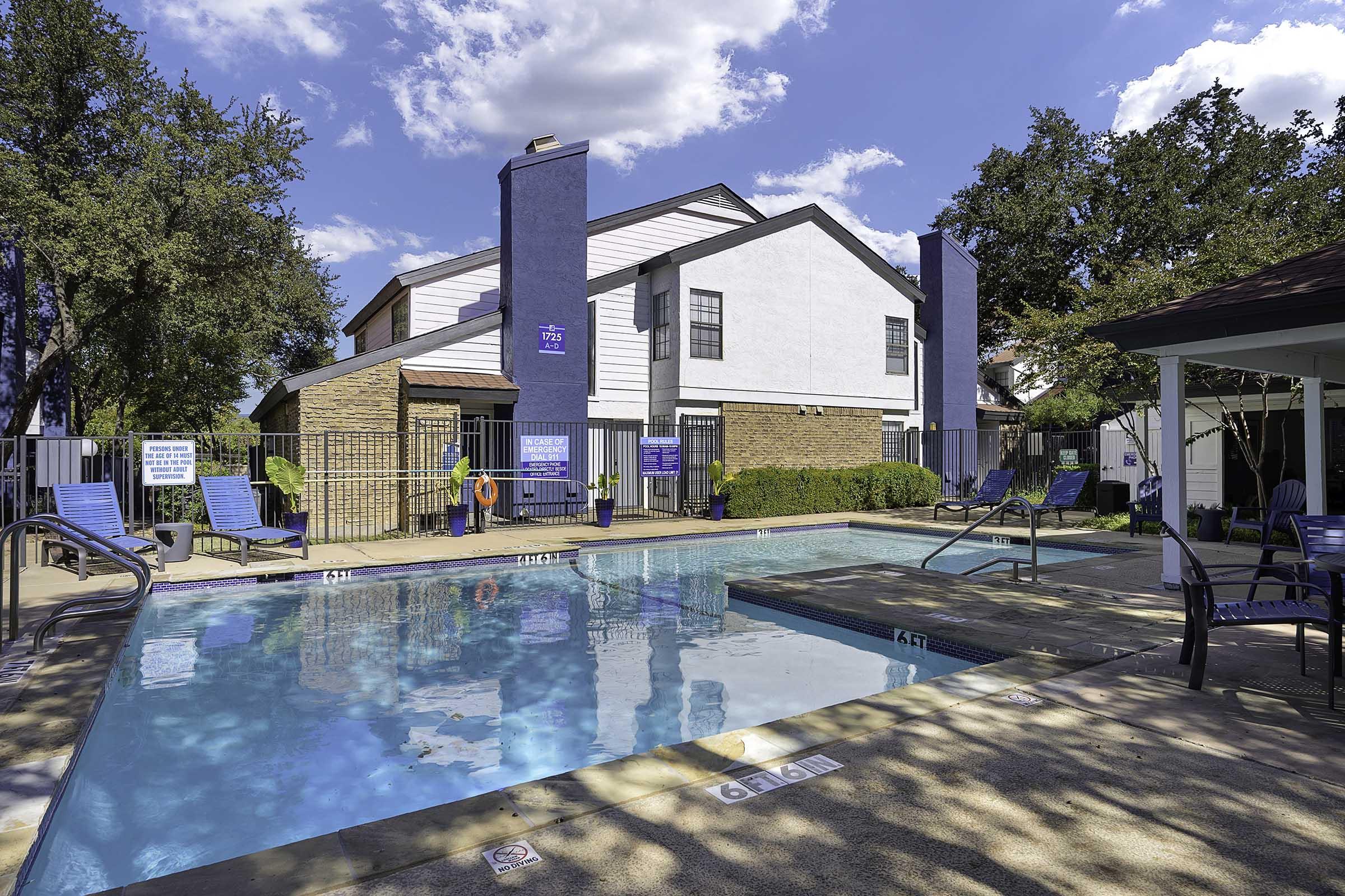 A clean, inviting swimming pool surrounded by lounge chairs and trees. The area features a brick and white building in the background, with blue accents on the pillars. Sunny skies and scattered clouds create a pleasant atmosphere, inviting relaxation and leisure.