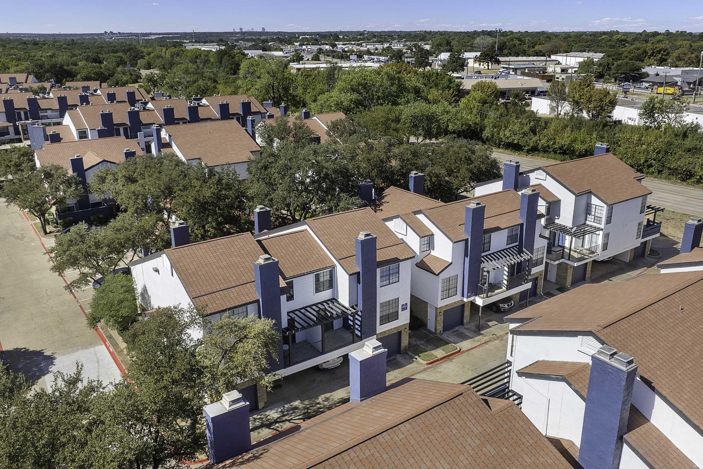 Aerial view of a residential complex featuring two-story buildings with red tiled roofs and blue accents. Surrounded by trees and parking spaces, the layout includes green areas and a nearby road visible in the background with distant city buildings.