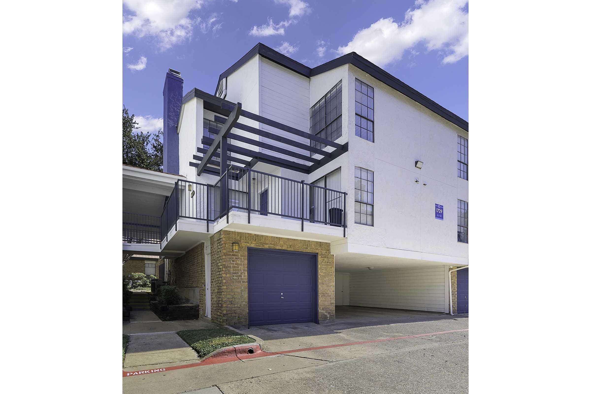 A modern multi-story building with a combination of white and brick exterior. The structure features large windows, a covered balcony with a black railing, and a garage below. The parking area is paved, and there is clear blue sky with a few clouds above.