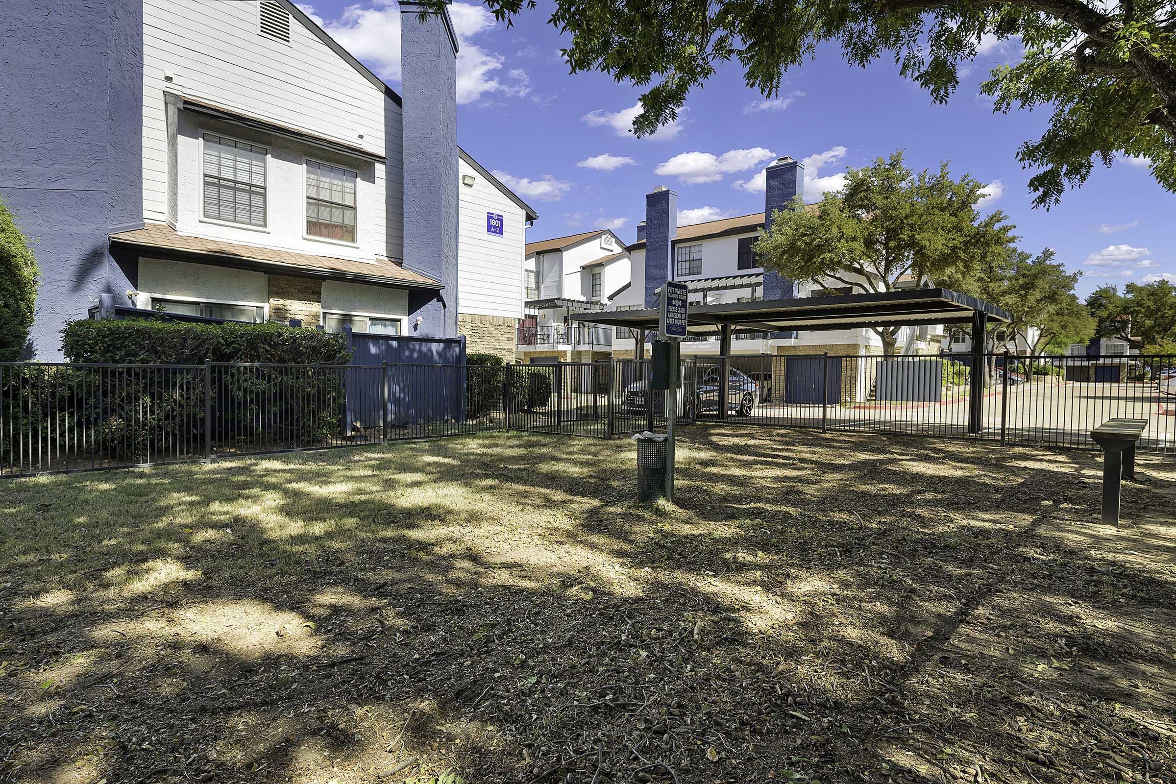 View of a grassy area with a fence, featuring a covered picnic area and trees. In the background, there are two multi-story apartment buildings with balconies. The sky is clear and blue, and there is a feeling of a well-maintained outdoor space suitable for relaxation or social gatherings.