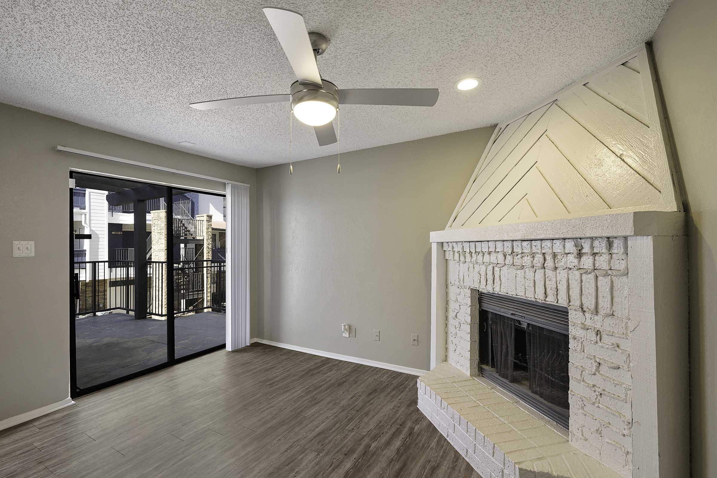 A modern living room featuring a ceiling fan and a decorative white brick fireplace. There is a sliding glass door leading to a balcony, allowing natural light to enter. The walls are painted in a neutral tone, and the floor has a wood laminate finish, creating a cozy and inviting atmosphere.
