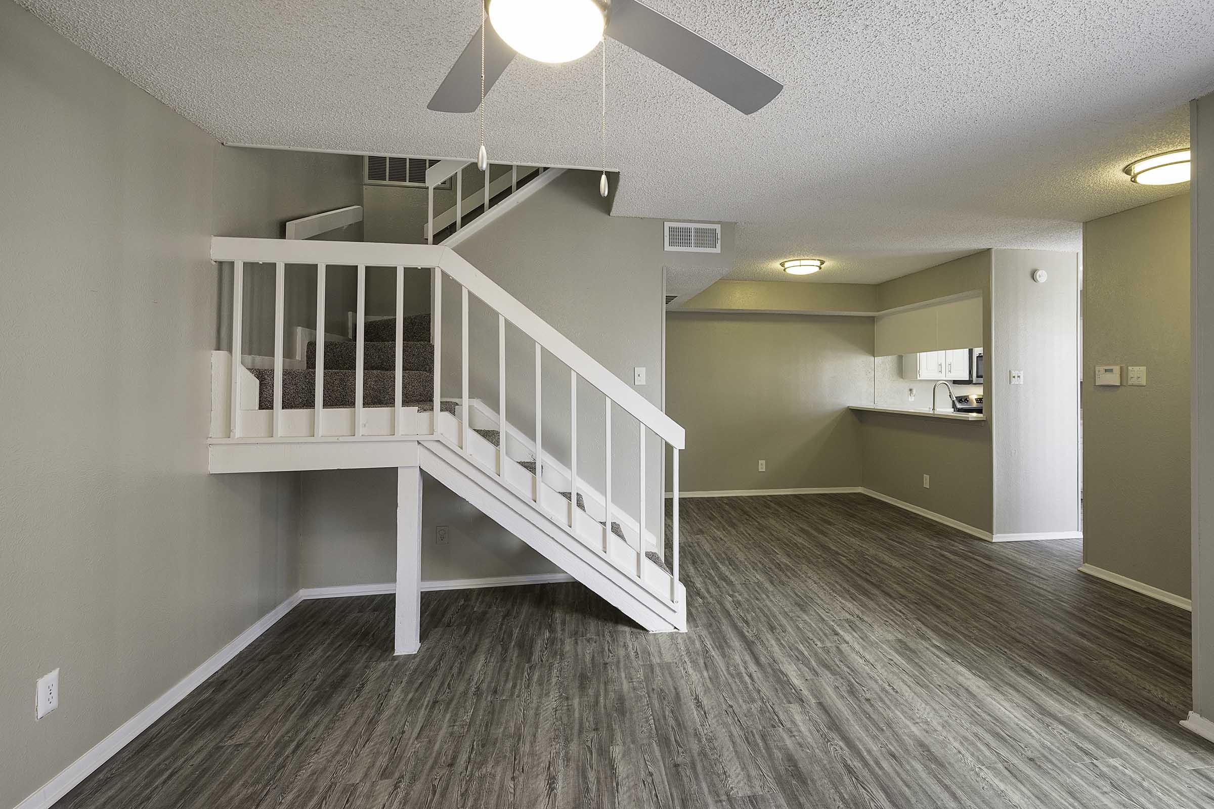 A spacious living area with a ceiling fan, featuring a staircase in the background leading to an upper level. The flooring is modern and wood-like, while the walls are painted in a light color. A kitchen area is visible in the distance, creating a welcoming atmosphere.