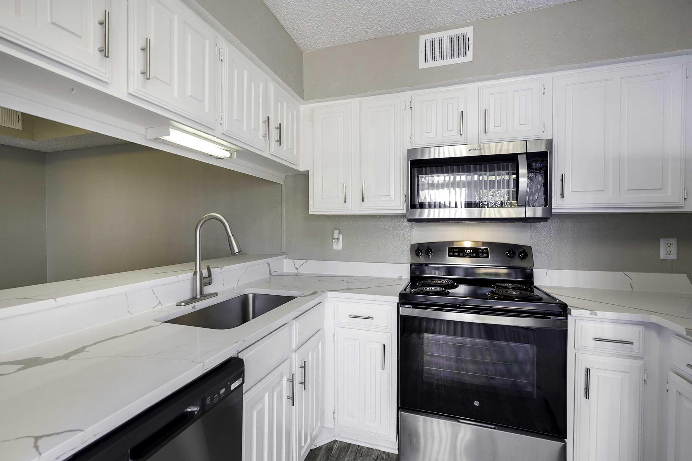 A modern kitchen featuring white cabinetry, a silver microwave, and a black stove. The countertops are light marbled, with a stainless steel sink and a black dishwasher visible. The space is well-lit and has a clean, contemporary look.
