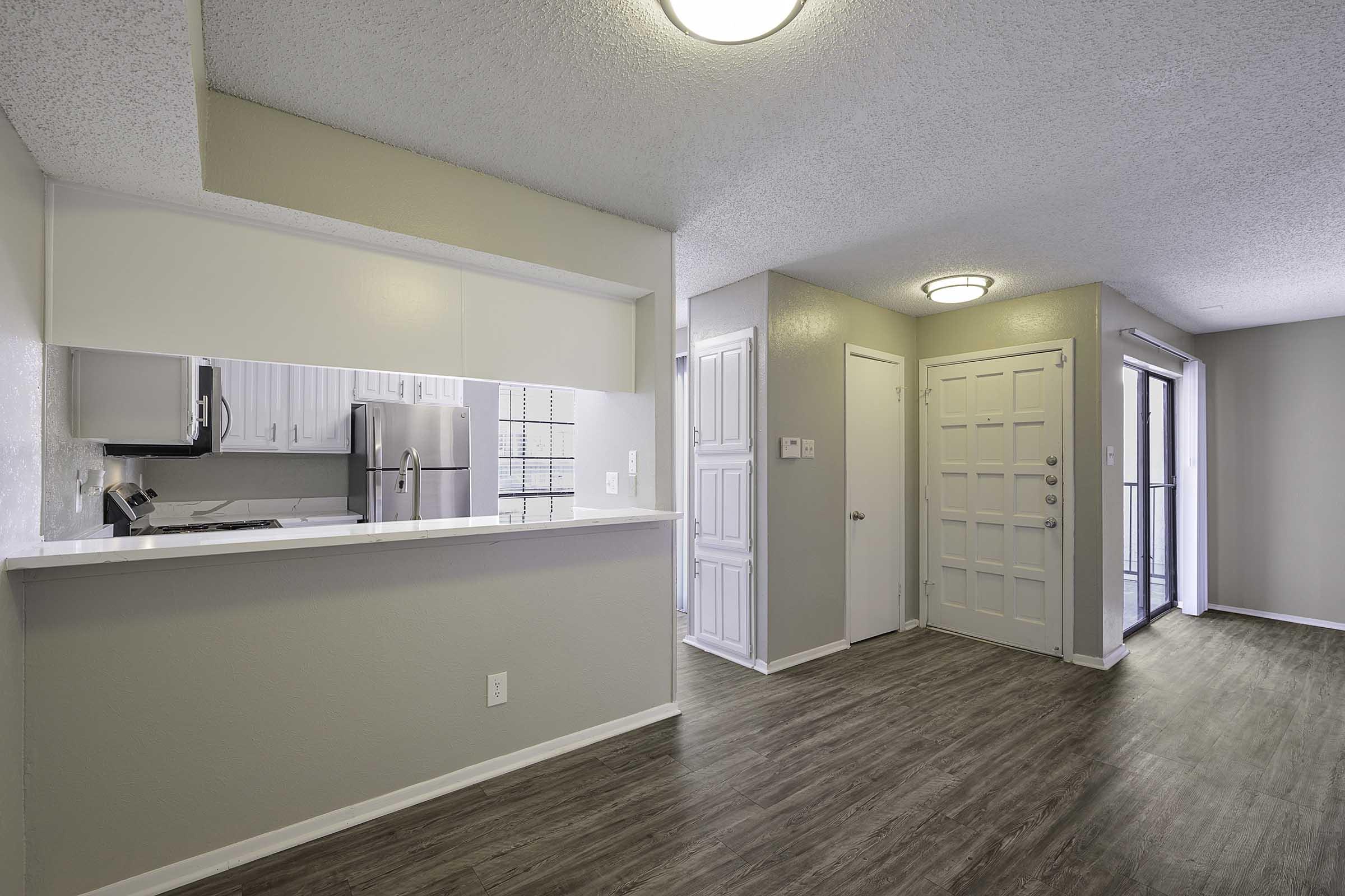 A modern kitchen view from the living area, featuring a bar counter, stainless steel appliances, and light-colored cabinets. The entrance door is visible with a bright interior, and sliding glass doors lead to an outdoor space. The floor is dark wood, creating a warm ambiance.
