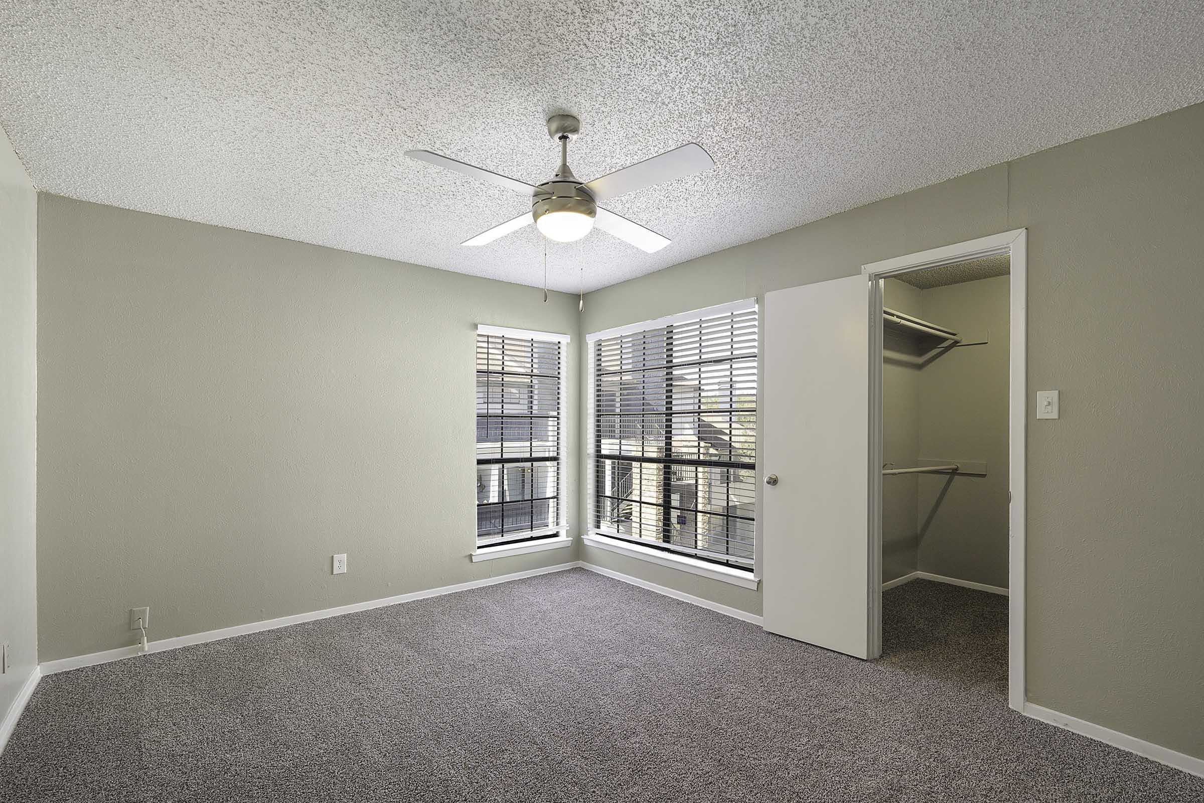 A well-lit, empty bedroom featuring light gray walls and carpet. Two windows with white blinds allow natural light, and a ceiling fan hangs from the textured ceiling. A closet is visible through an open door on the right, with neutral decor creating a cozy atmosphere.