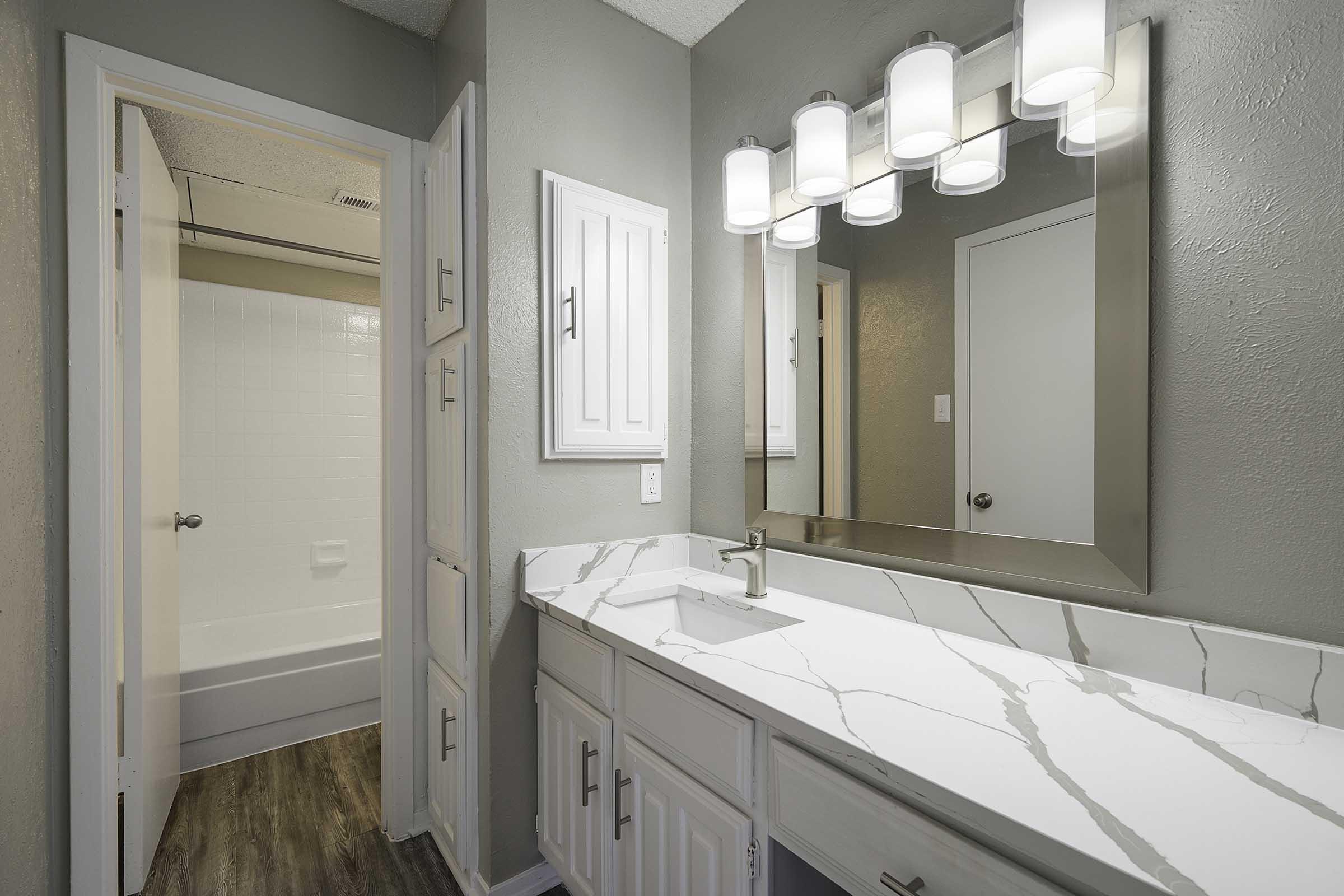 A modern bathroom interior featuring a marble countertop with a sink, illuminated by a vanity light fixture above. A mirror is mounted on the wall, and there is a doorway leading to a tub area in the background. The walls are painted gray, and the floor has a wood-like appearance.