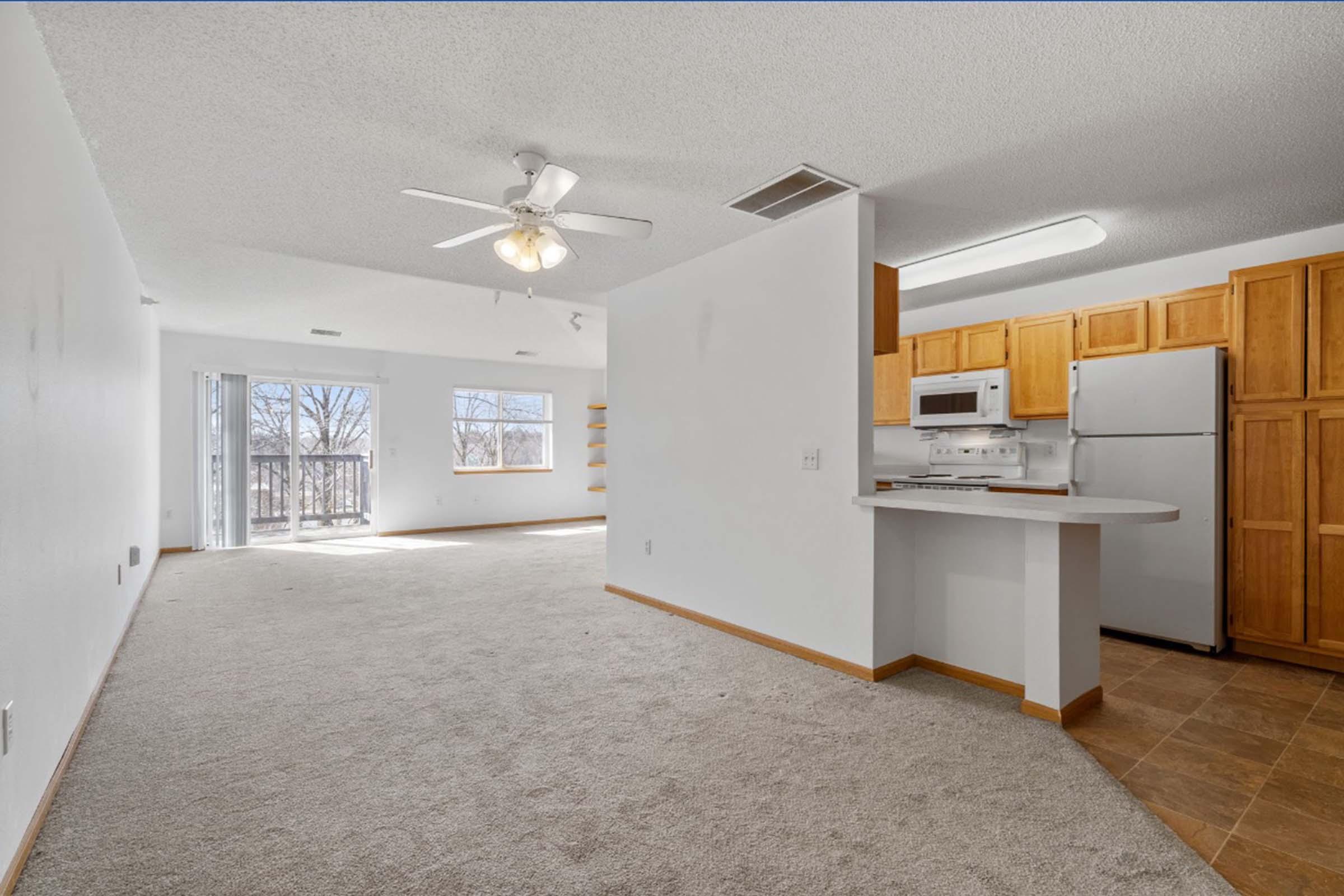 A spacious, well-lit living area featuring light-colored carpet, a ceiling fan, and a set of sliding glass doors leading to a balcony. The kitchen is open to the living space, equipped with wooden cabinets, a white microwave, and a refrigerator against the wall. Natural light fills the room through two windows.