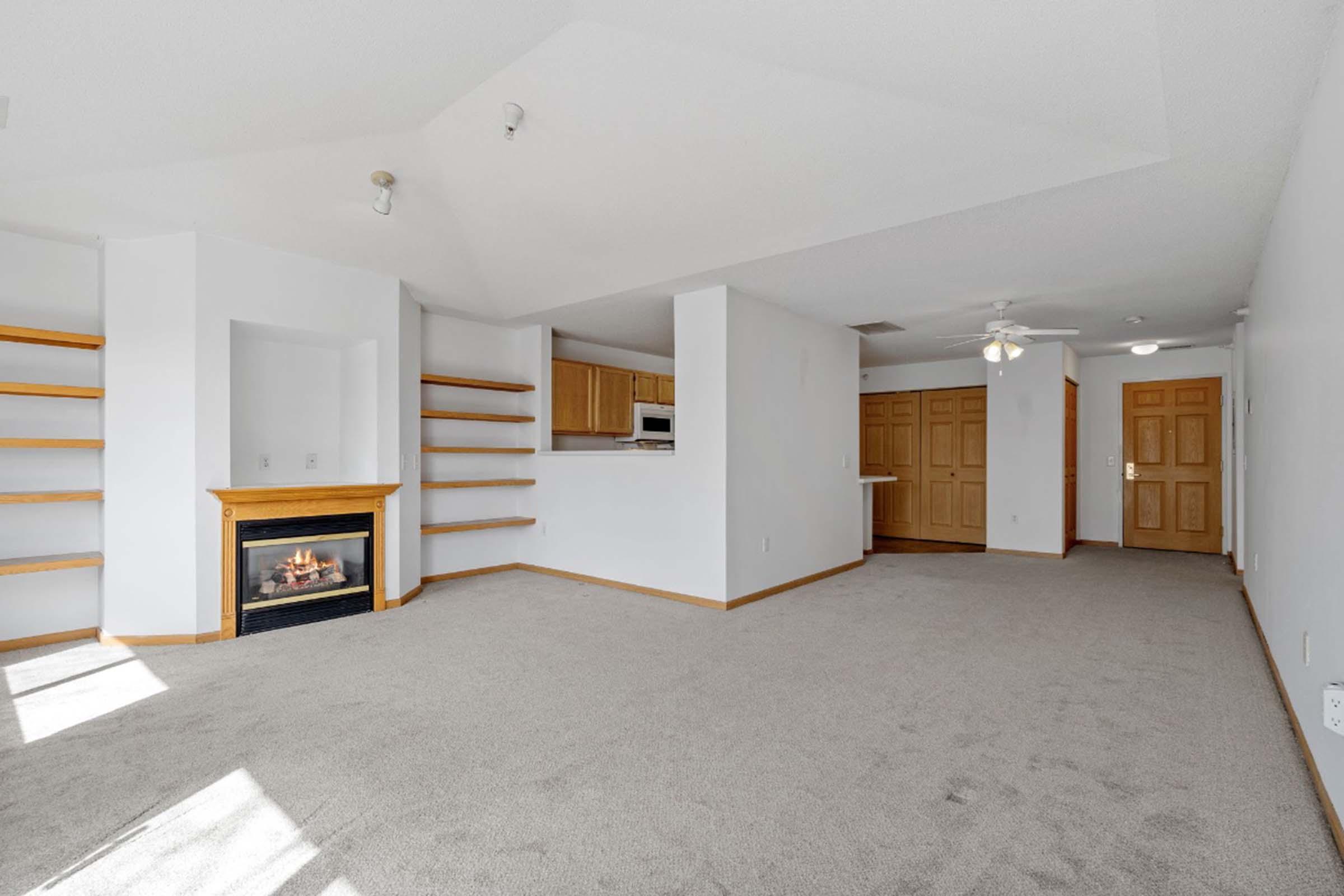 Spacious and bright living area featuring plush carpeting, a cozy fireplace, and built-in shelves. The open layout connects to a kitchen area with a breakfast bar, and light-colored walls create an inviting atmosphere. Two doorways lead to the main entrance and other rooms.