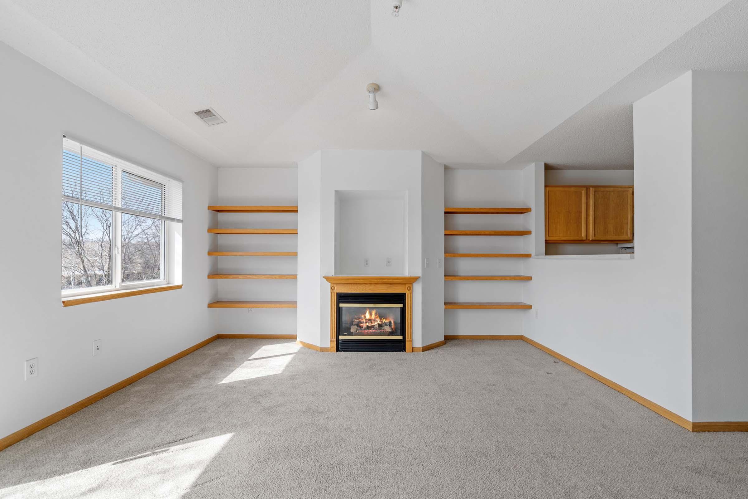 A bright, empty room with beige carpeting and white walls. There are large windows letting in natural light, and a modern fireplace in the center with wooden shelves on either side. The ceiling is sloped, adding to the spacious feel of the room.
