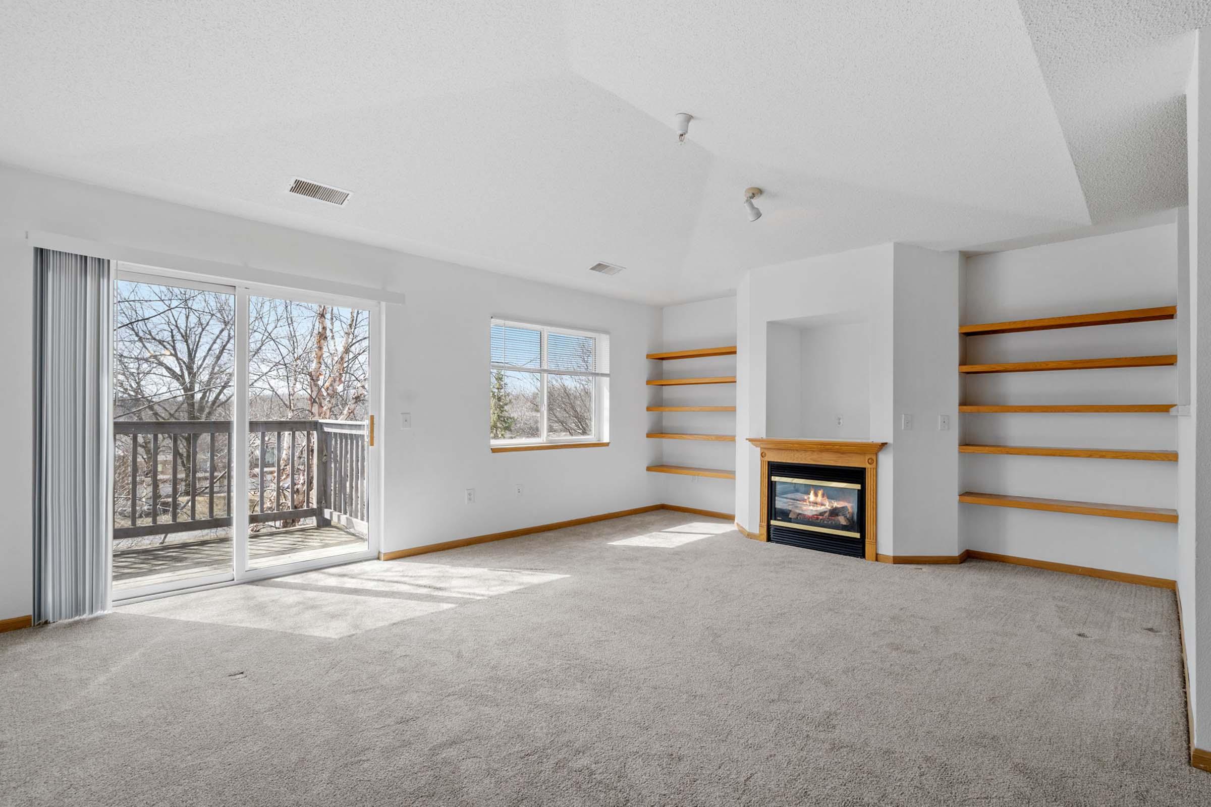 A spacious living room featuring a cozy fireplace, large windows that bring in natural light, and built-in shelves. There's a sliding door leading to a balcony, with a neutral carpet and walls, creating a bright and inviting atmosphere.
