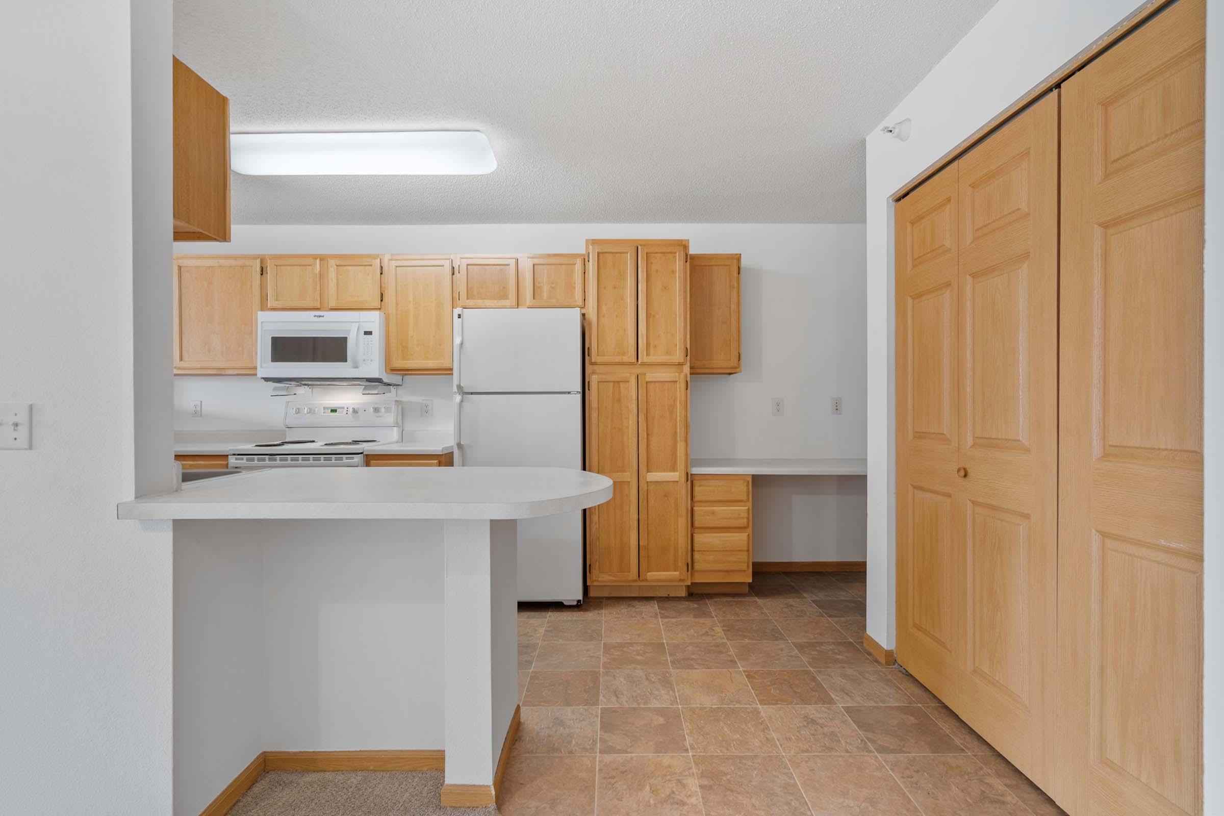 A modern kitchen with light wood cabinetry, a white refrigerator, and a microwave above the stove. The space features beige tile flooring and a breakfast bar that separates the kitchen from a small nook with built-in shelves. Two large closet doors are visible, suggesting ample storage.