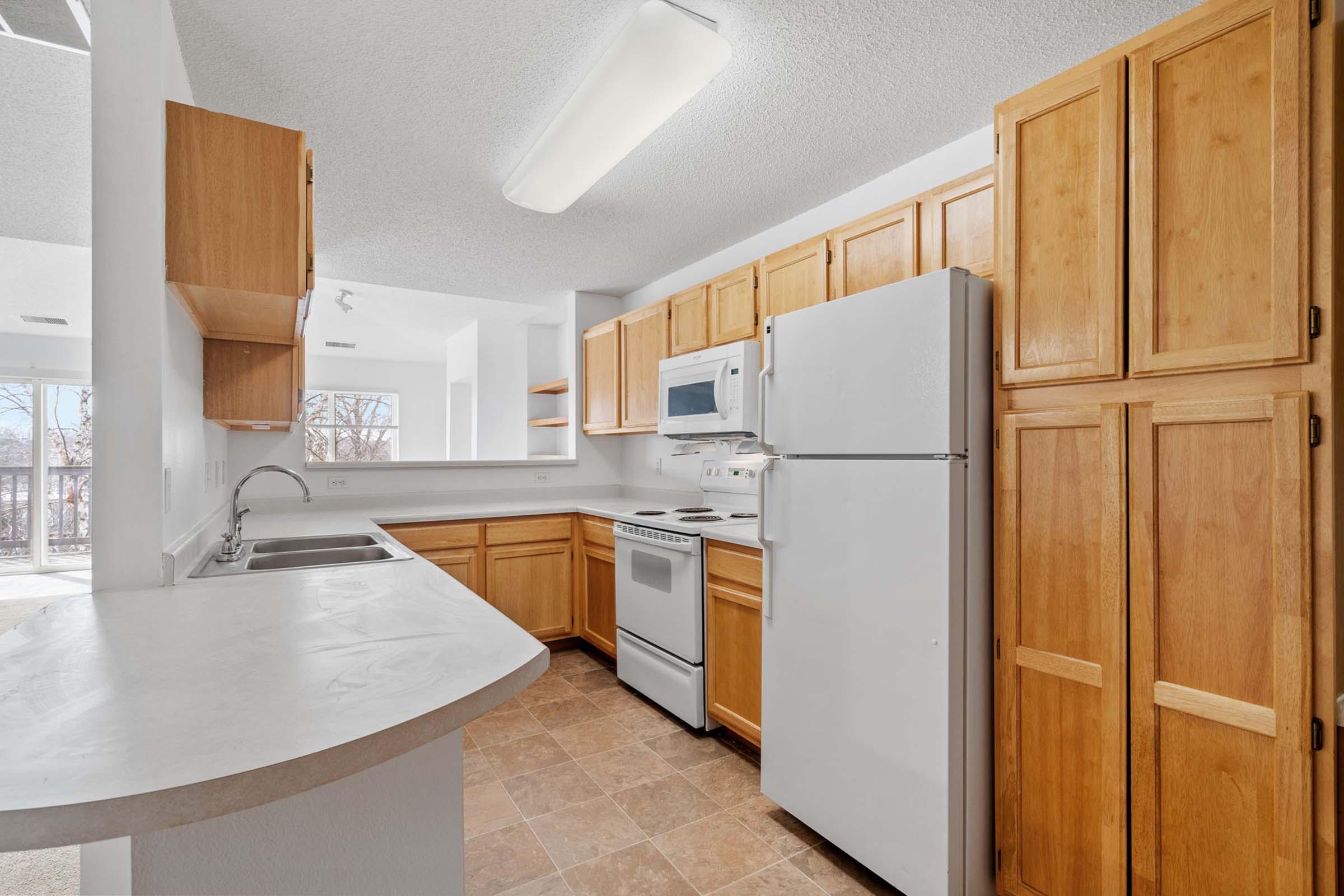 A bright kitchen featuring wooden cabinets, a white refrigerator, and a microwave. The countertop is light-colored with a sink. The kitchen has a stove and oven, with a window providing natural light, and an open layout connecting to a living area. The floor is tiled with a light design.