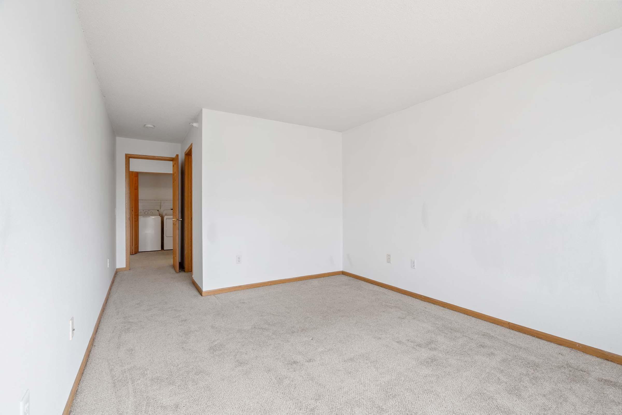Spacious empty room with light gray carpet and white walls, featuring wooden door frames. Visible entrance leads to a laundry area in the background. Natural light brightens the space, creating an airy feel.
