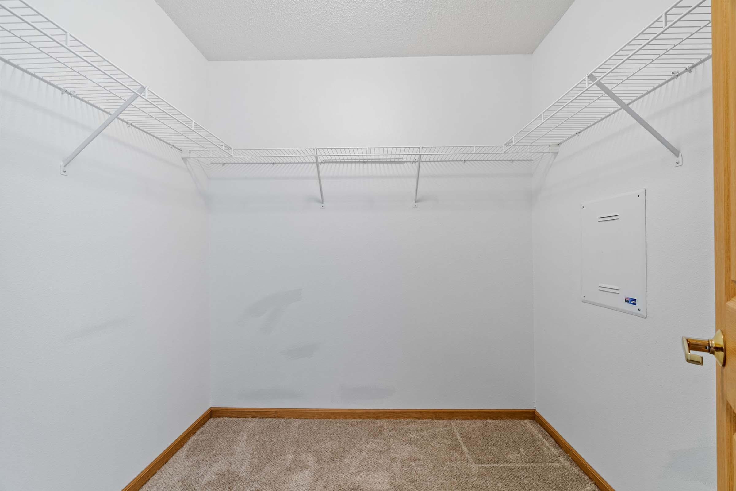 Empty closet with white walls and a tan carpet. The closet features a wire shelving unit along the back wall and has a small electrical panel on the right side. A wooden door is partially visible on the right, leading into the room. The overall appearance is clean and uncluttered.