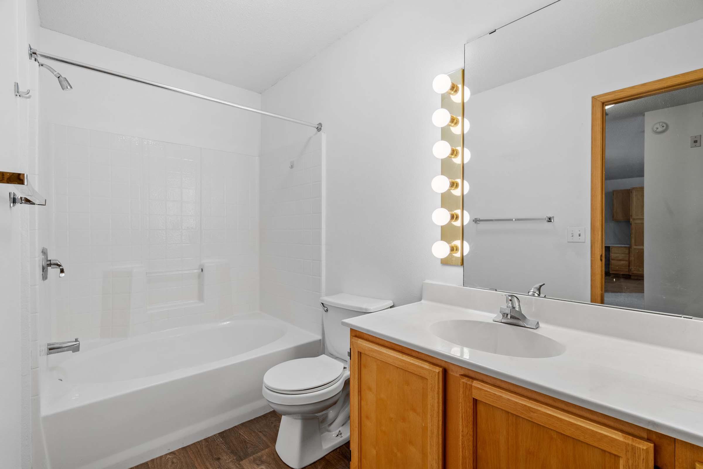 Bright and clean bathroom featuring a bathtub with a shower, a white tiled wall, and a large mirror illuminated by six round vanity lights. The vanity has wooden cabinetry and a modern sink. The flooring is a natural wood style, creating a warm atmosphere.