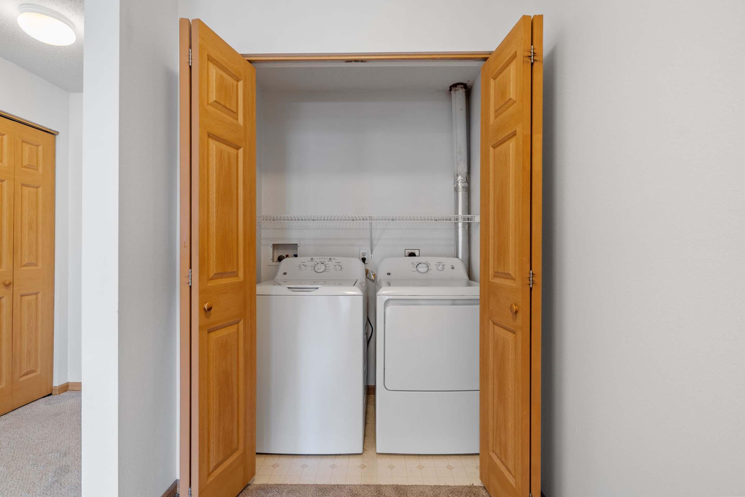A well-lit laundry room featuring a front-loading washing machine and a dryer side by side, enclosed by double wooden doors. The walls are light-colored, and there's a shelf above the appliances. A glimpse of a carpeted hallway is visible in the foreground.