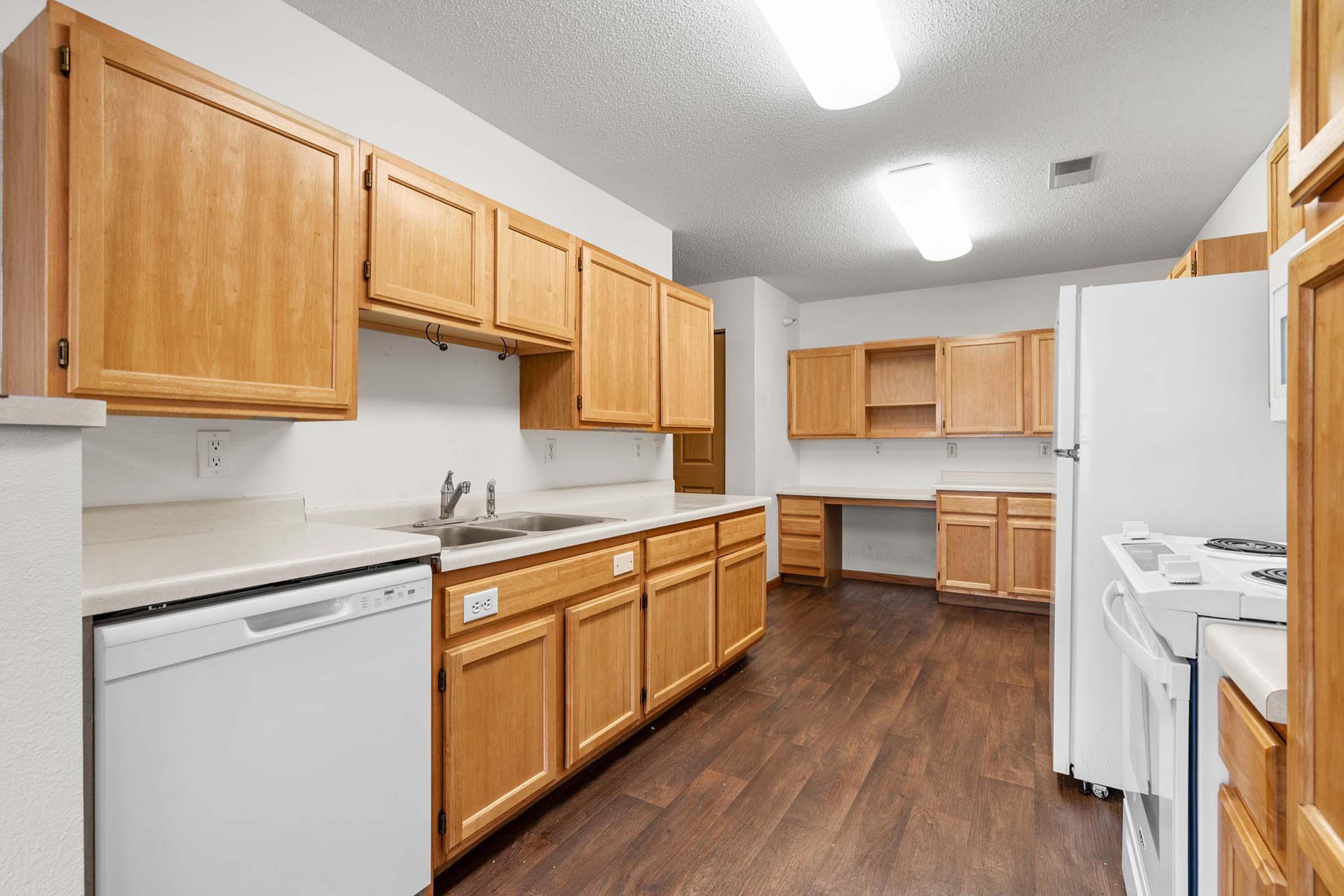 A spacious kitchen featuring light wooden cabinets, a double sink, a white dishwasher, and a white refrigerator. The room has a clean, modern layout with ample counter space and laminate flooring, illuminated by overhead lighting.