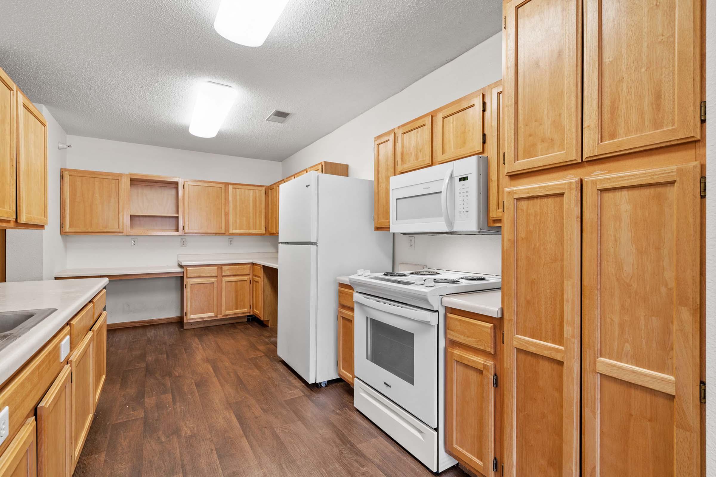 A spacious kitchen featuring light wood cabinetry, a white refrigerator, microwave, and stove. The room has a modern laminate floor and overhead fluorescent lighting, providing a bright and functional cooking space.