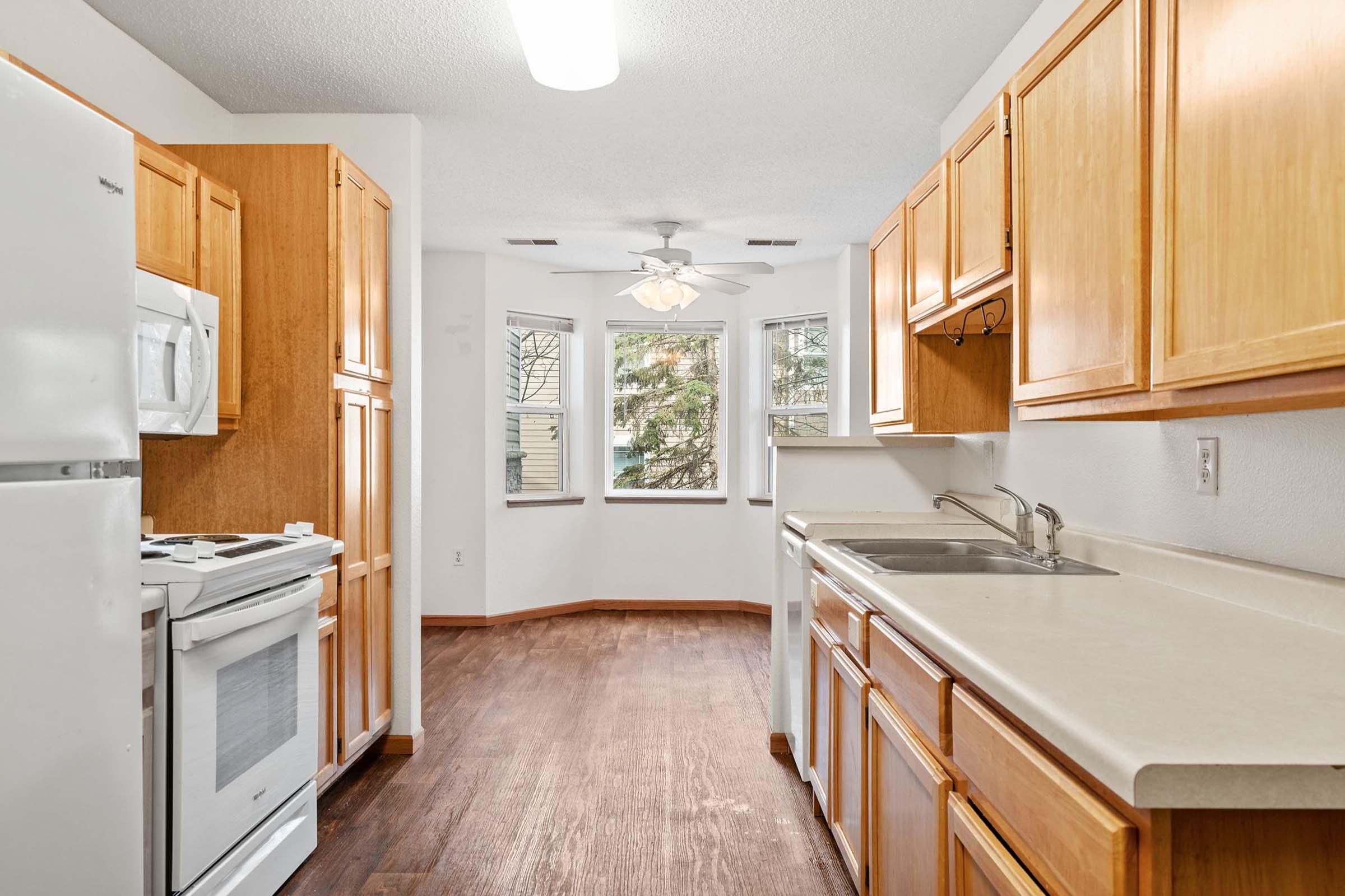 A bright kitchen featuring wooden cabinets, a white stove, and a refrigerator. The countertop is light-colored, with a sink beneath a window. Natural light enters through adjacent windows, and the flooring is dark wood. A ceiling fan adds to the airy atmosphere.